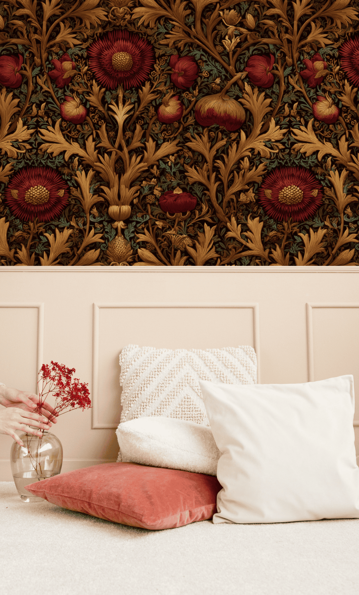 A hand places red dried flowers in a glass vase beside white and terracotta floor cushions, set against a wall adorned with Baroque Bloom Wallpaper’s ornate floral motifs and beige paneling for a timeless vibe.