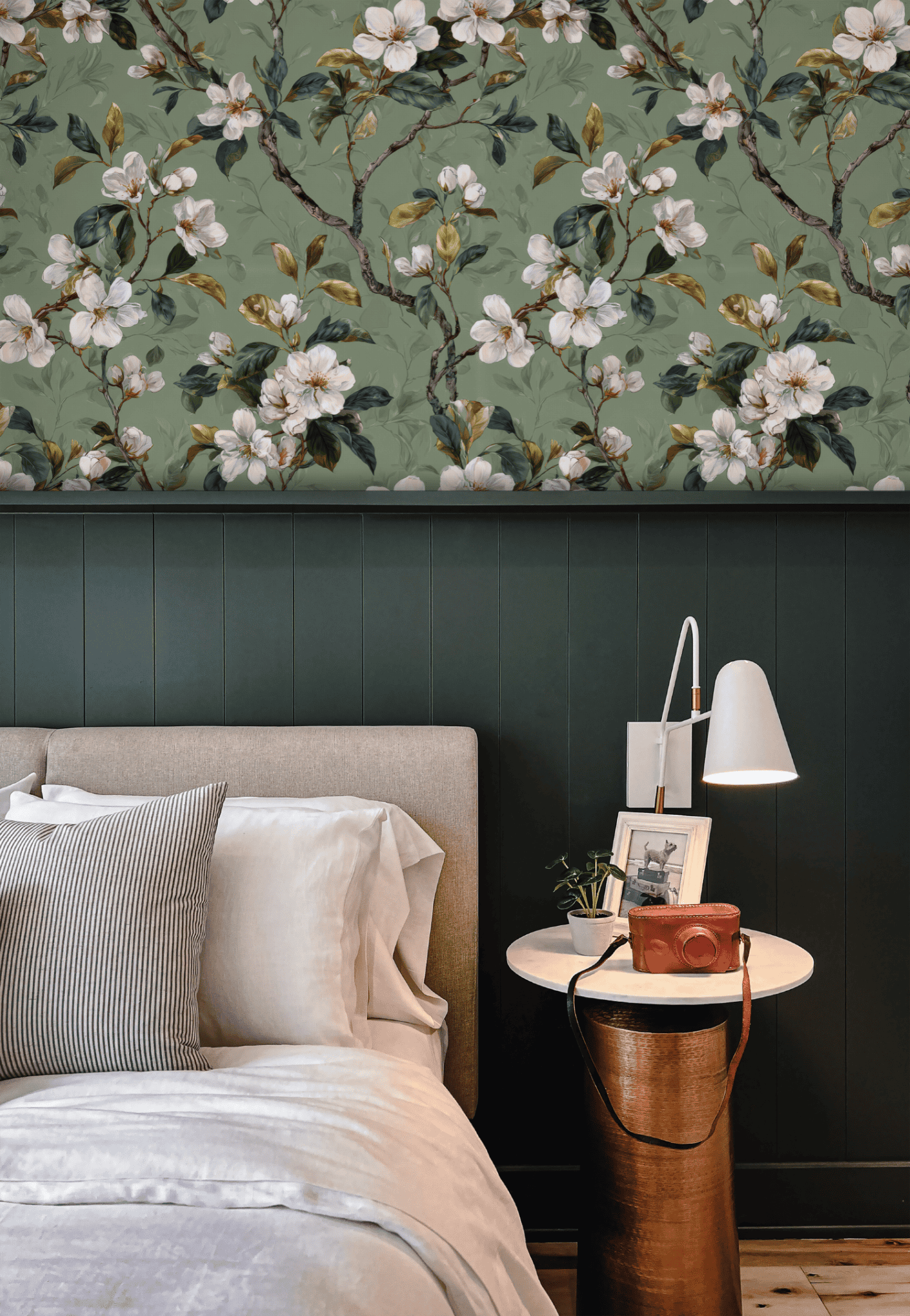 A modern bedroom with a light upholstered bed, striped pillows, a round side table holding books, a camera, and a white lamp, set against elegant green floral Blossom Serenity Wallpaper and a dark paneled wall.