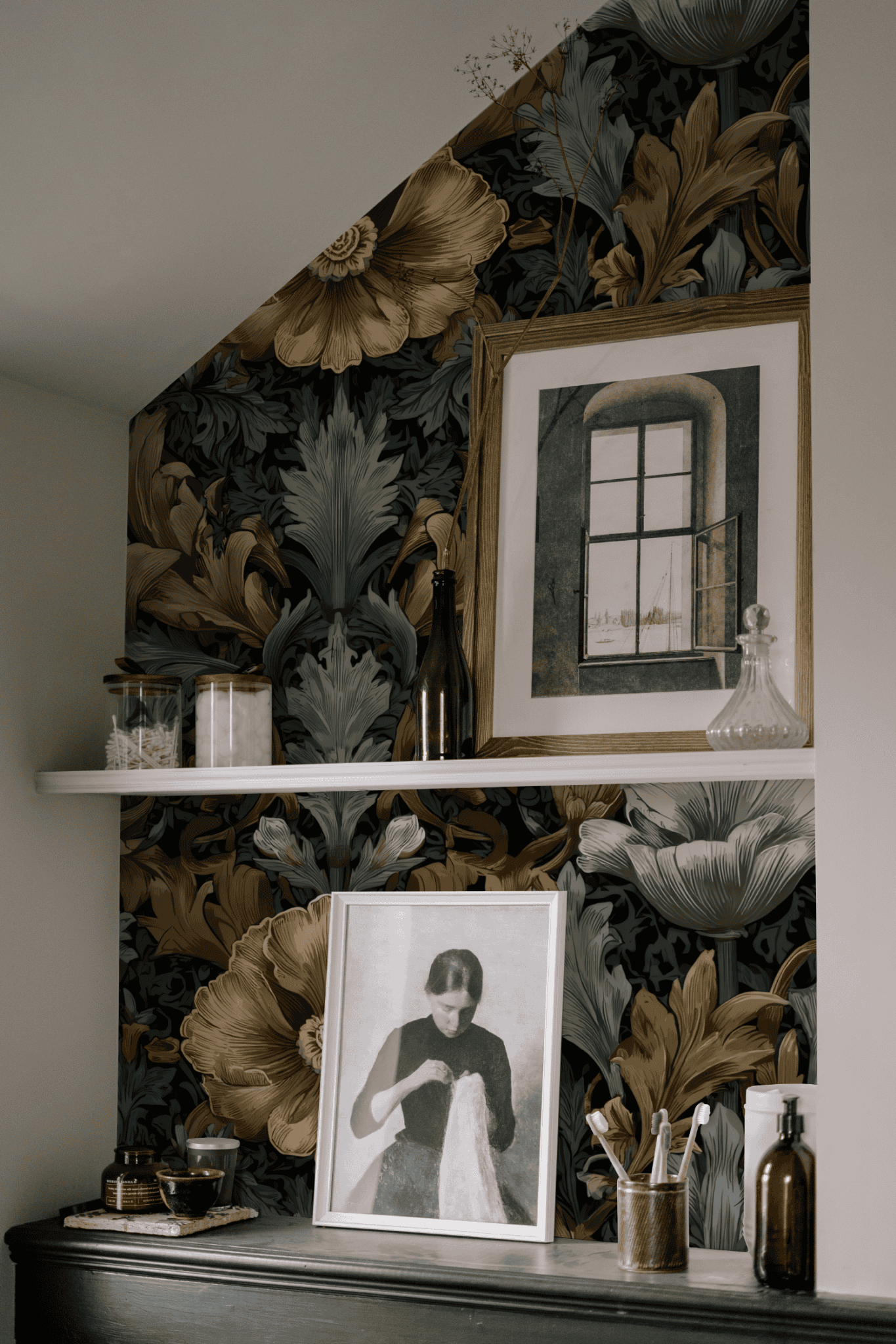 A wall adorned with Midnight Blooms Wallpaper exudes timeless elegance, featuring two framed pictures, glass jars, and decorative bottles arranged on a white shelf and a black surface below.