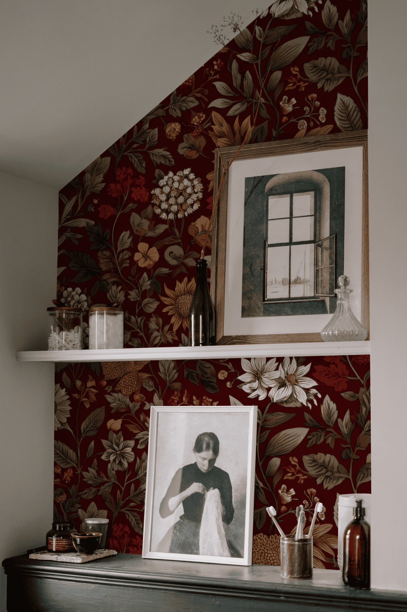 Against the Crimson Bloom Wallpaper, a shelf holds bottles, jars, and framed art, featuring a woman’s portrait and a window illustration.