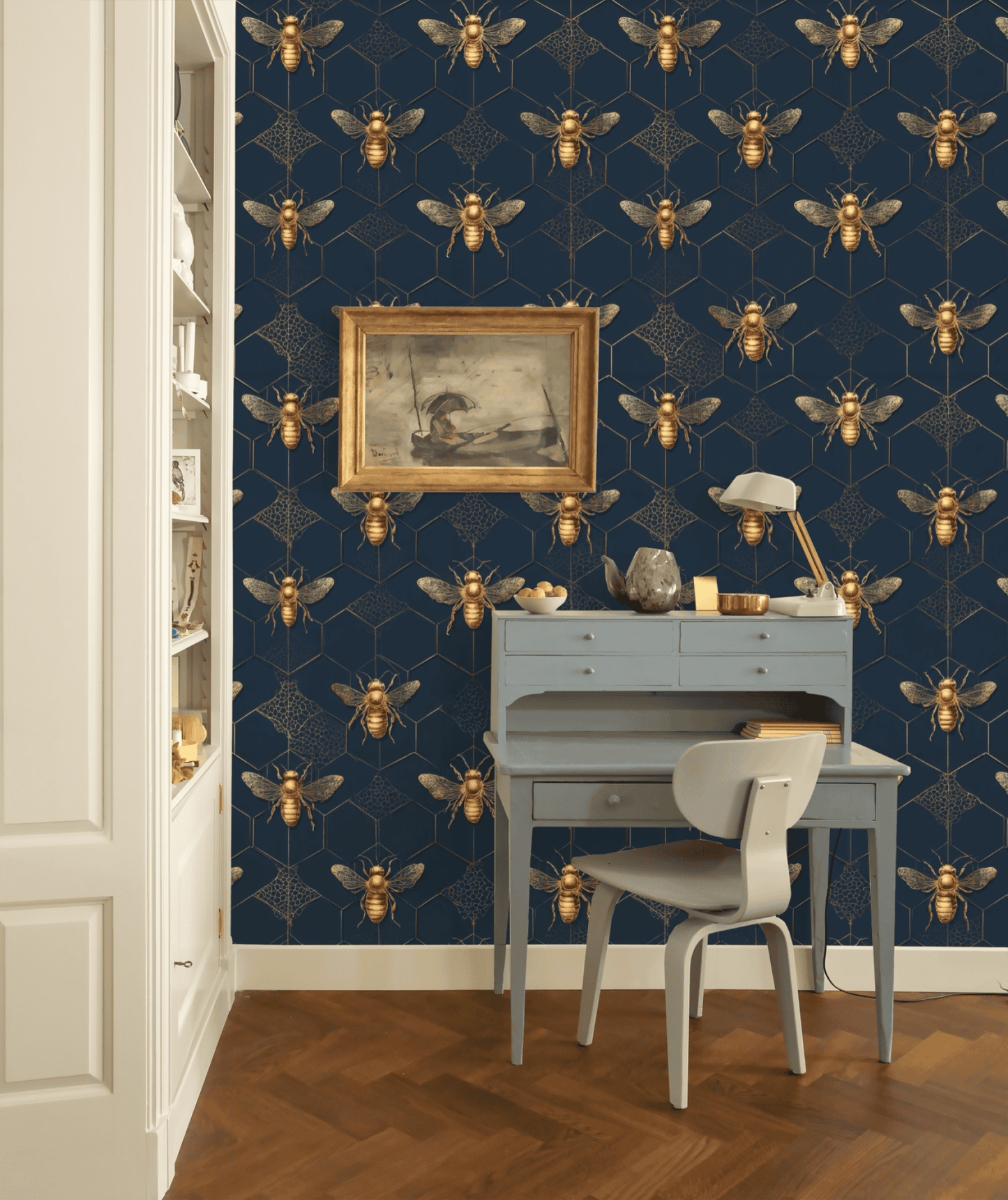 A small blue desk and chair sit against a wall covered in Golden Hive Wallpaper; a framed painting and desk lamp rest on the desk.