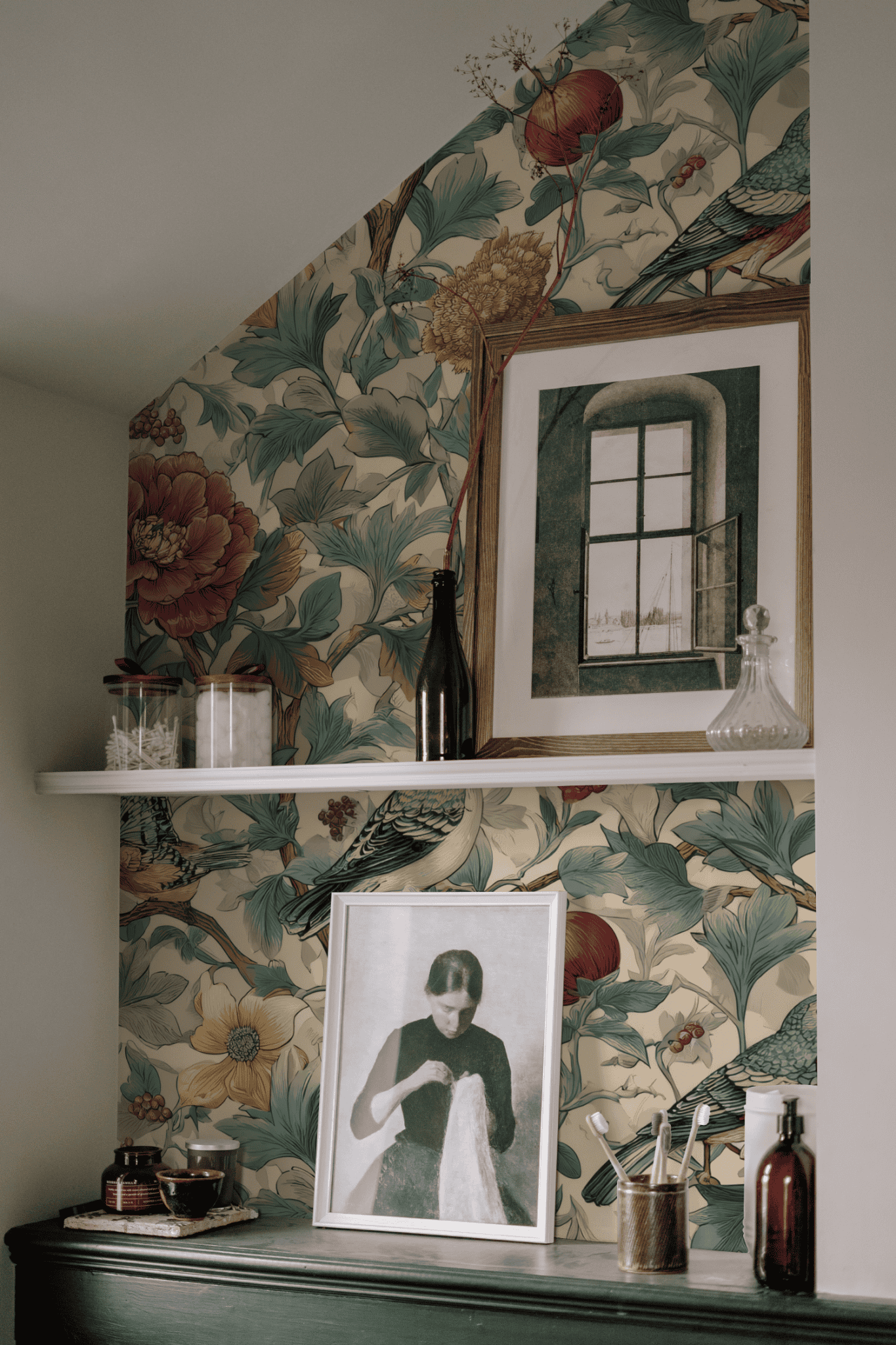 A shelf with framed art, glass bottles, and jars is set against a wall featuring Aviary Bloom Wallpaper with lush florals and vibrant birds. A black-and-white portrait photo sits on the mantel below.