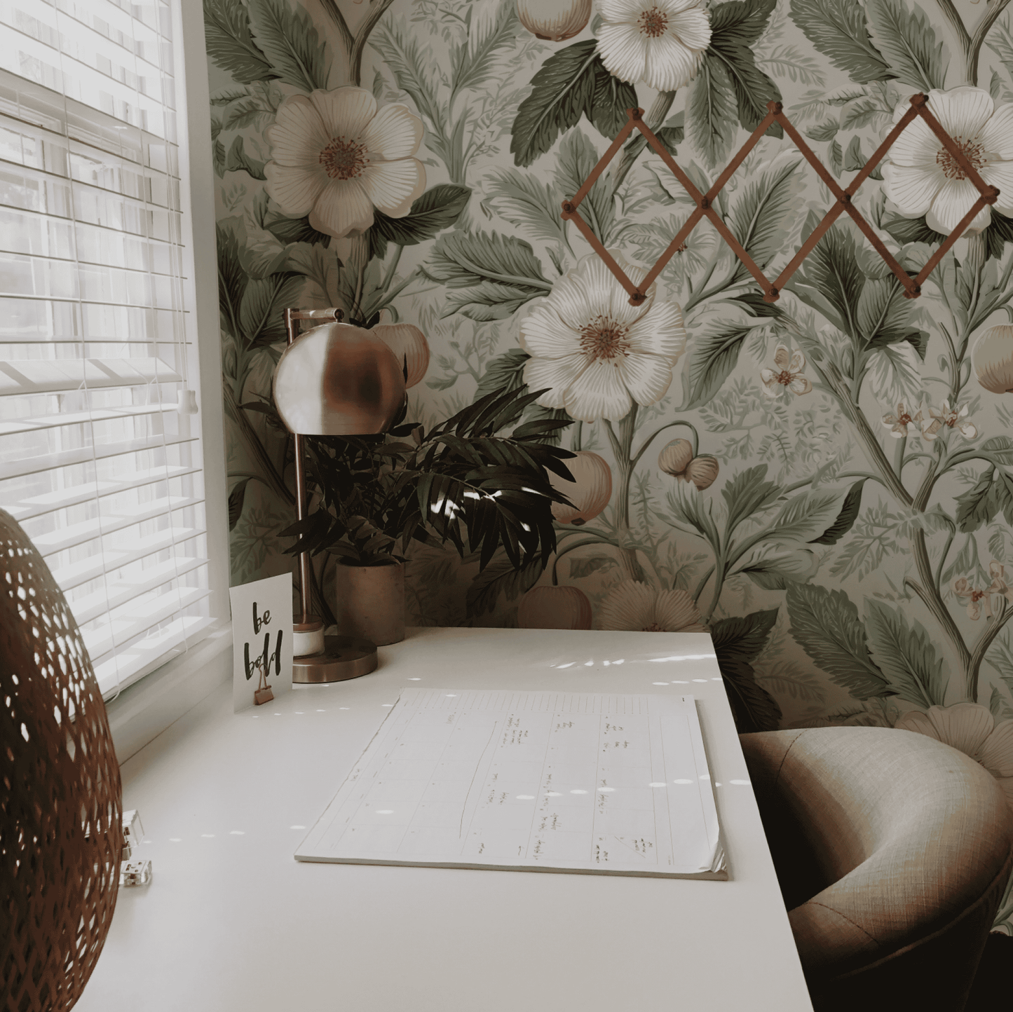 A white desk with a calendar, lamp, plant, and “be bold” card sits by a window; the wall features Eden's Bloom Wallpaper for botanical charm, complemented by a wooden accordion rack.