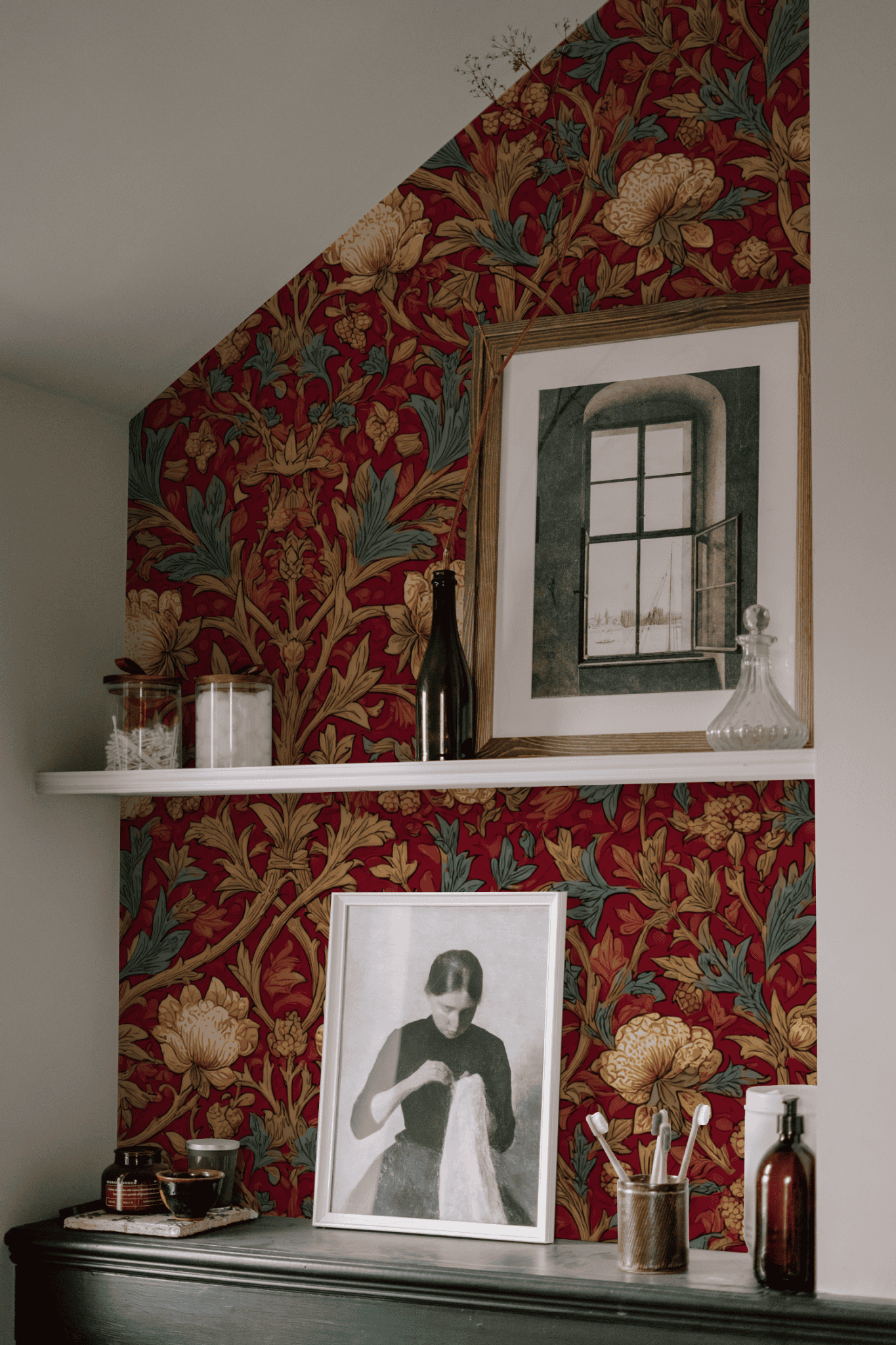 A shelf set against Crimson Garden Wallpaper displays glass bottles, jars, and framed art—a photo of a woman and a painting of a window.