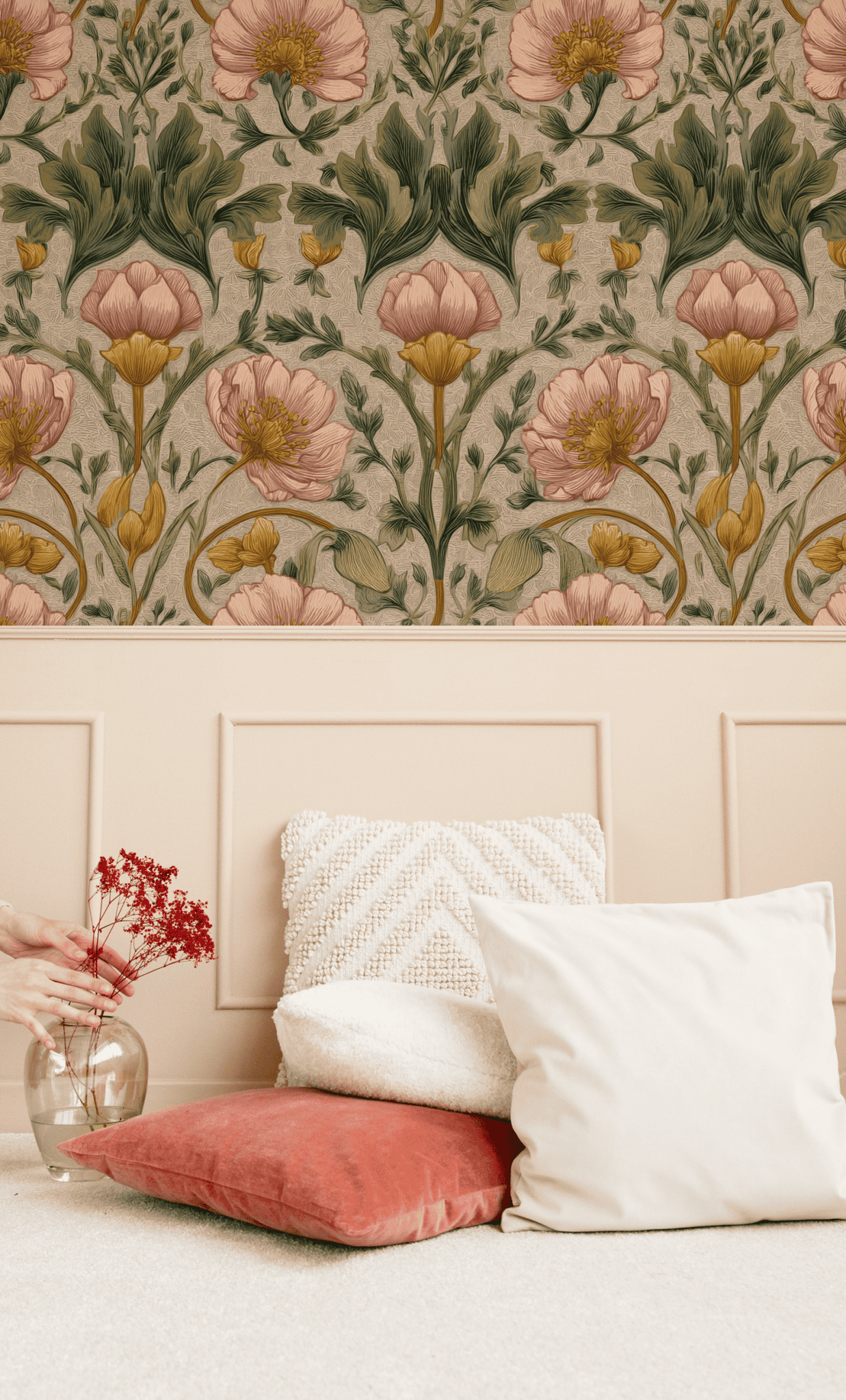 Three cushions sit on the floor beside a wainscoted wall adorned with Floral Elegance Wallpaper, while a hand holds a vase of delicate flowers, creating a scene of timeless beauty.