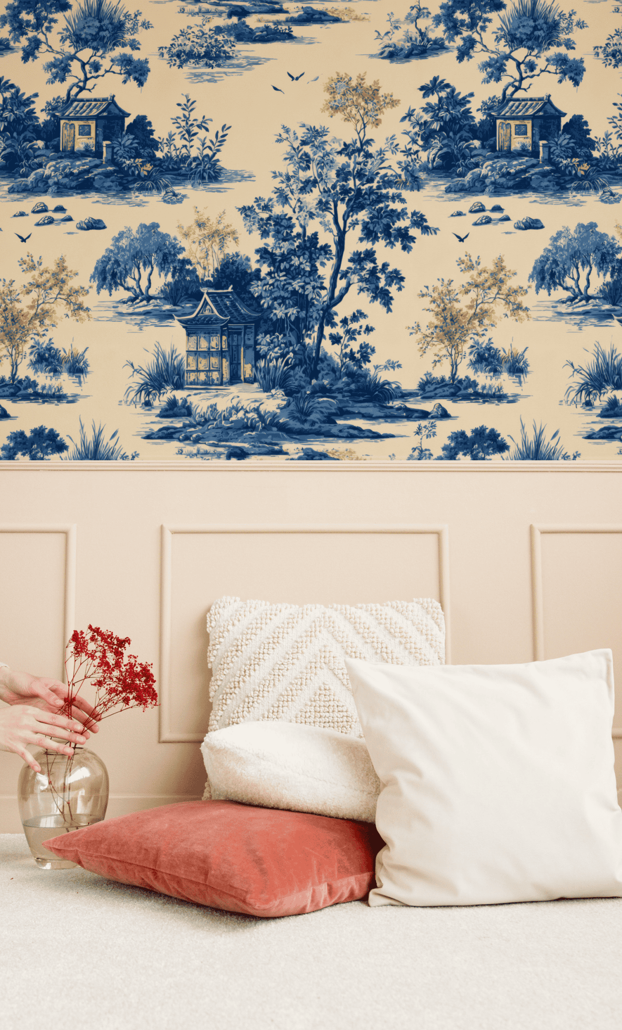 A hand places a glass vase with red flowers beside white and coral pillows on a cream carpet, set against the timeless elegance of Azure Serenity Wallpaper’s blue and beige Asian toile designs.