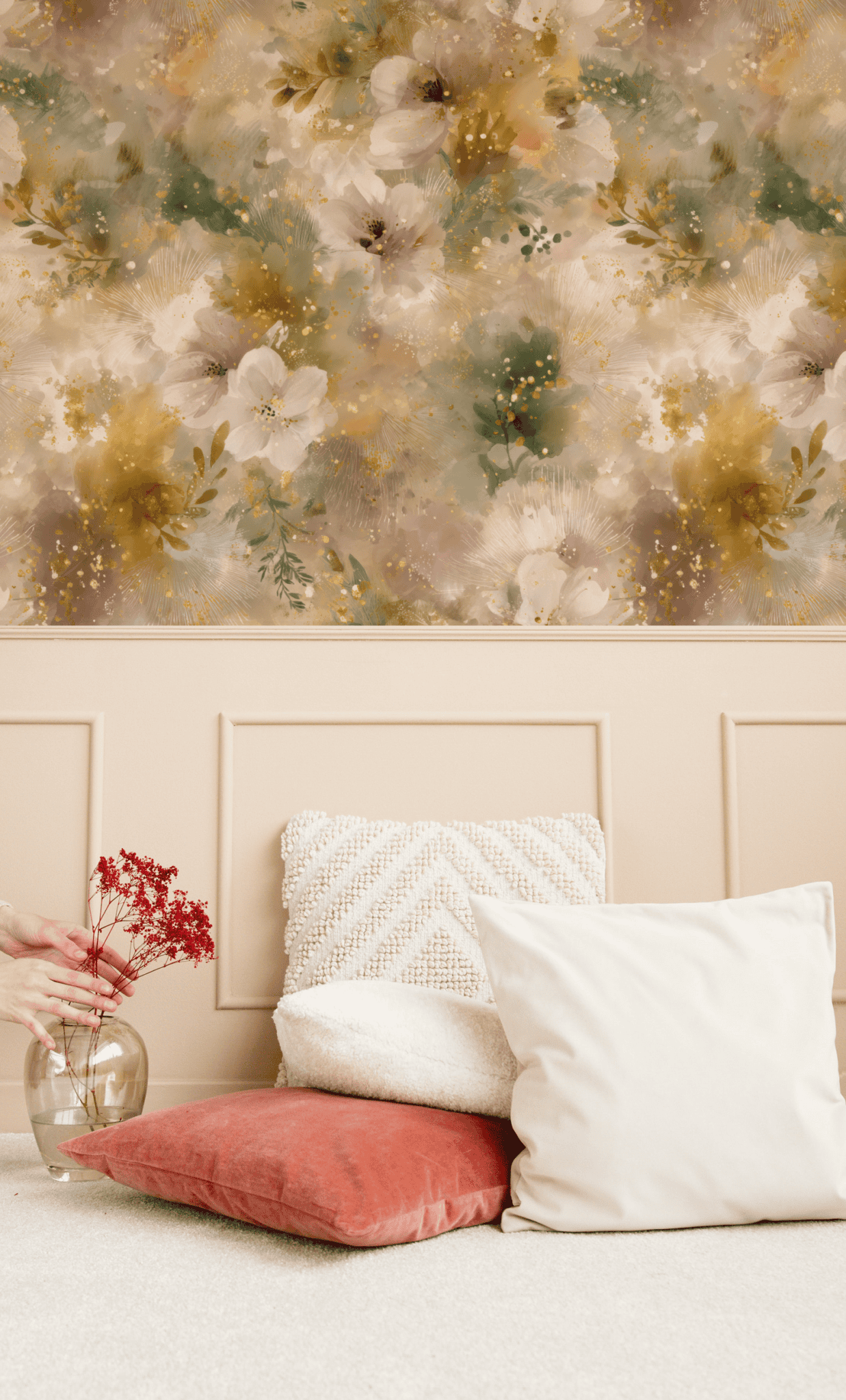 A person places red flowers in a clear vase on the floor beside beige and rust pillows, with Whispering Blooms Wallpaper featuring floral swirls and light paneling in the background.