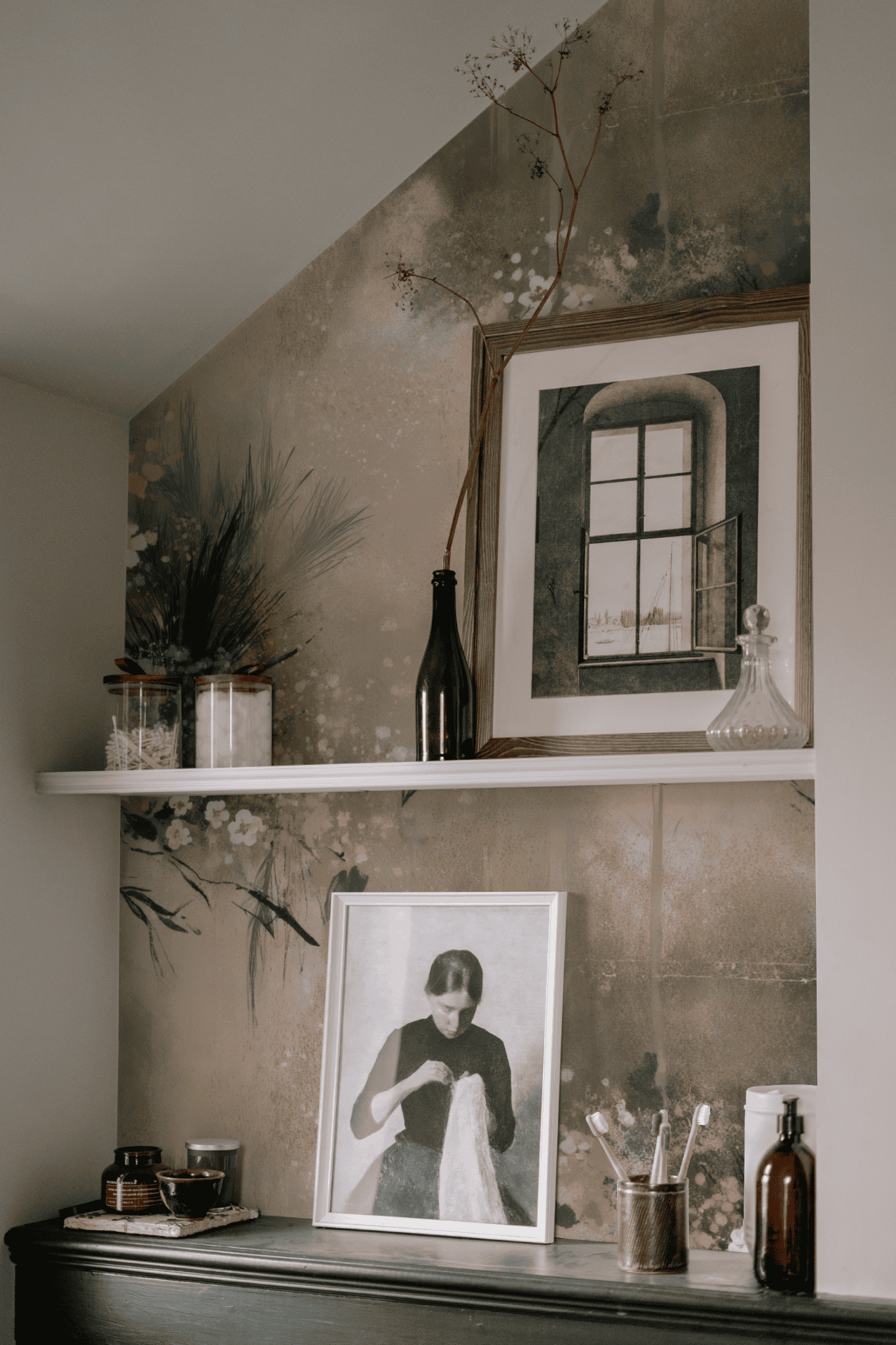 A shelf displays framed art, dried plants in a bottle, glassware, and small containers; Serene Blossom Wallpaper’s pastel floral design and soft hues add dreamy serenity while lower art shows a woman holding fabric.
