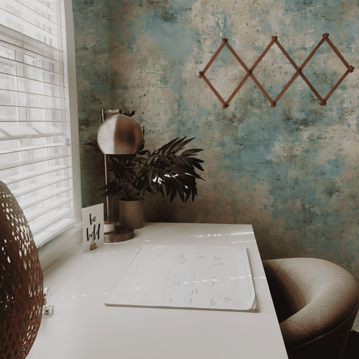 A minimalist home office desk with a lamp, plant, calendar, and 'be bold' card sits by a window with blinds; the Tranquil Mist Wallpaper in soft blue tones and an empty geometric rack create a calming ambiance.