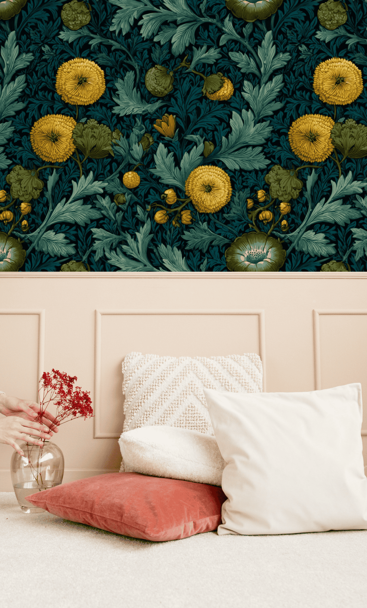 A decorative wall with Golden Flora Wallpaper featuring dark florals and teal foliage above a beige panel, matched with beige and coral cushions, as a hand arranges red flowers in a glass vase on a light carpeted floor.