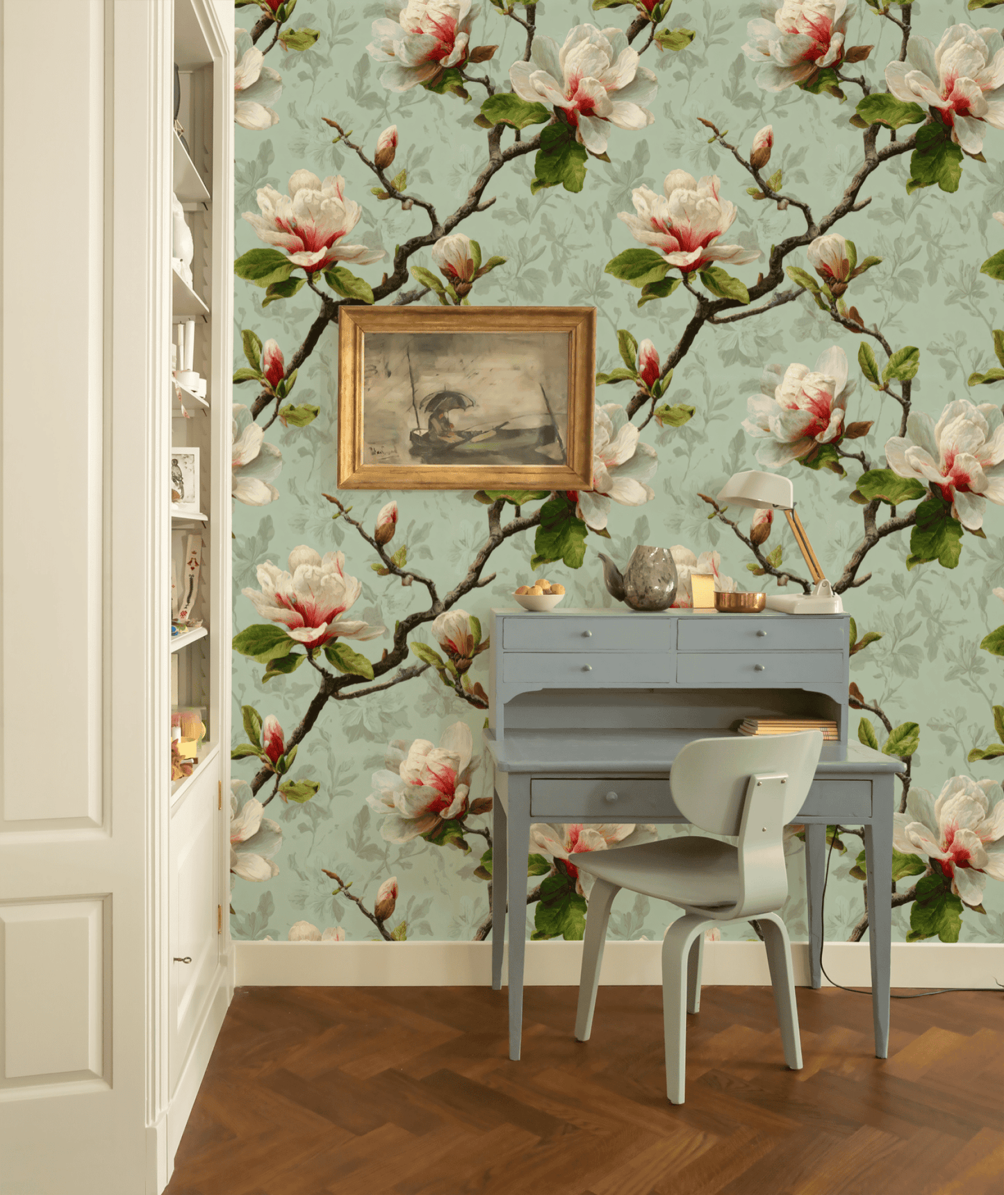 A small writing desk with chair sits against Magnolia Blossom Wallpaper, topped by a framed bird painting and flanked by shelves of books and decor, creating a gentle botanical atmosphere.