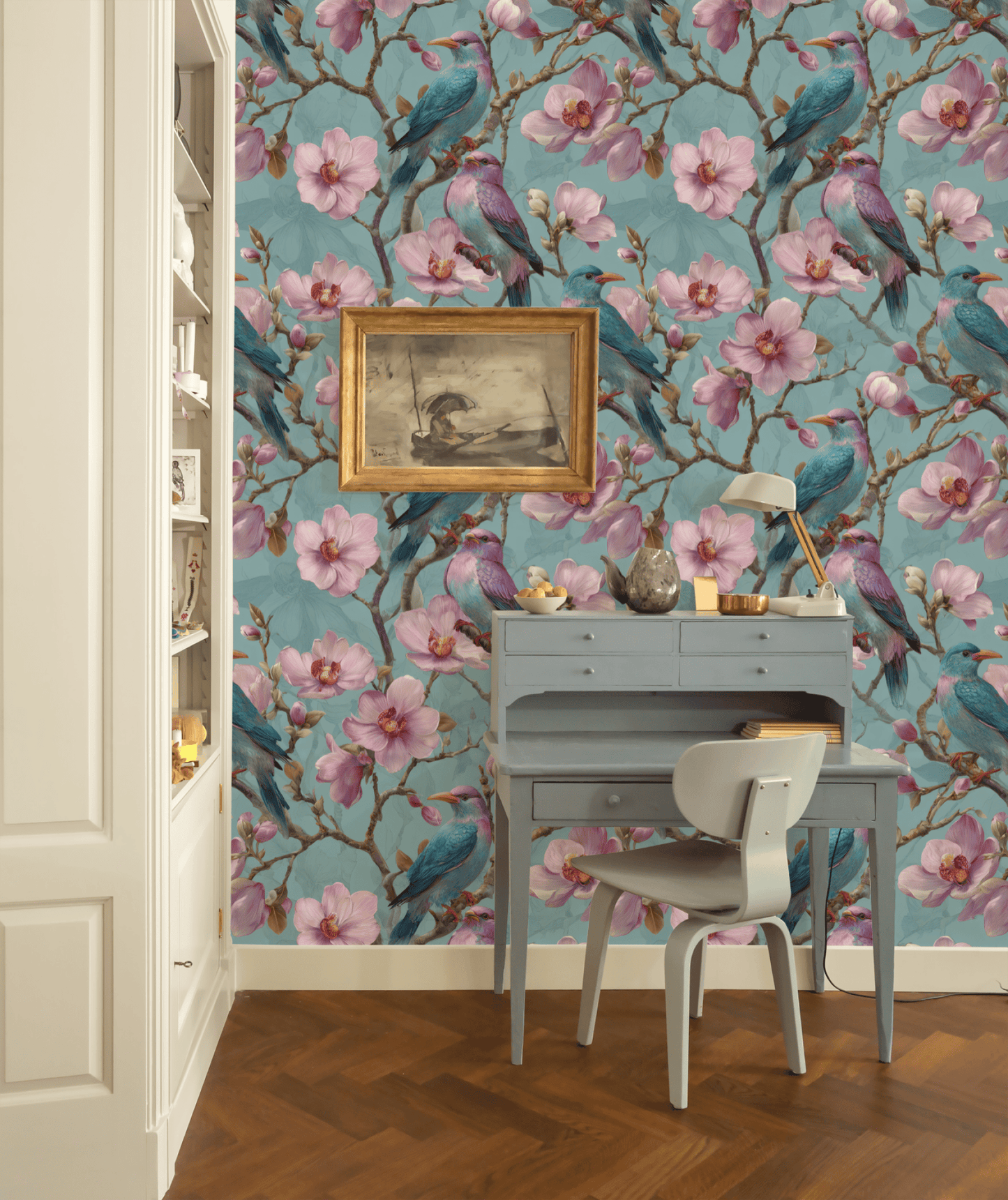 A blue desk and chair stand before a teal wall covered in Blossom Aviary Wallpaper, adorned with vibrant birds and blooming flowers; above the desk hangs a framed bird artwork.