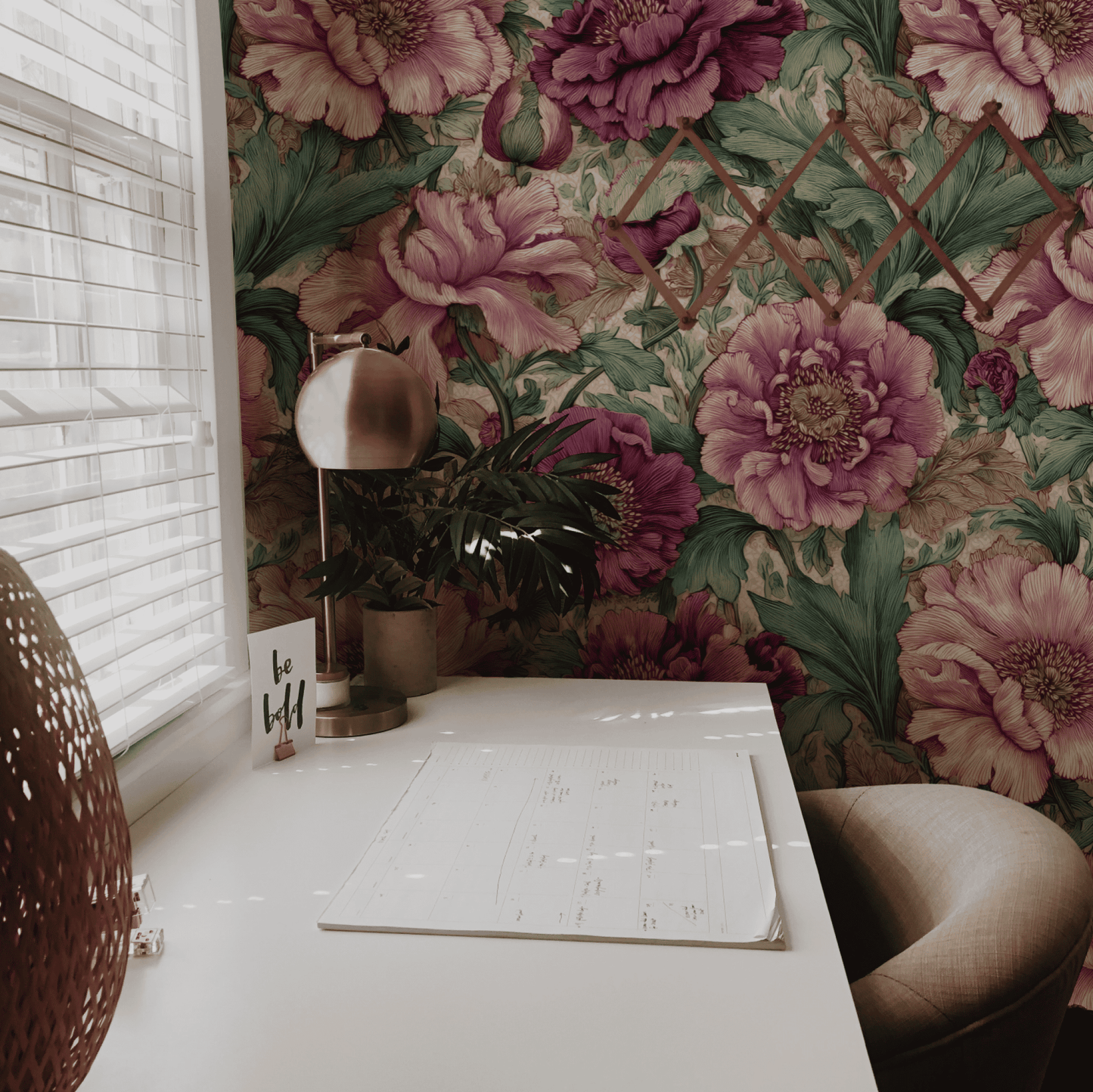 A white desk with a lamp, plant, and calendar sits by a window with blinds; the wall is adorned with Peony Elegance Wallpaper, featuring soft pink peonies and green leaves for a sophisticated atmosphere.