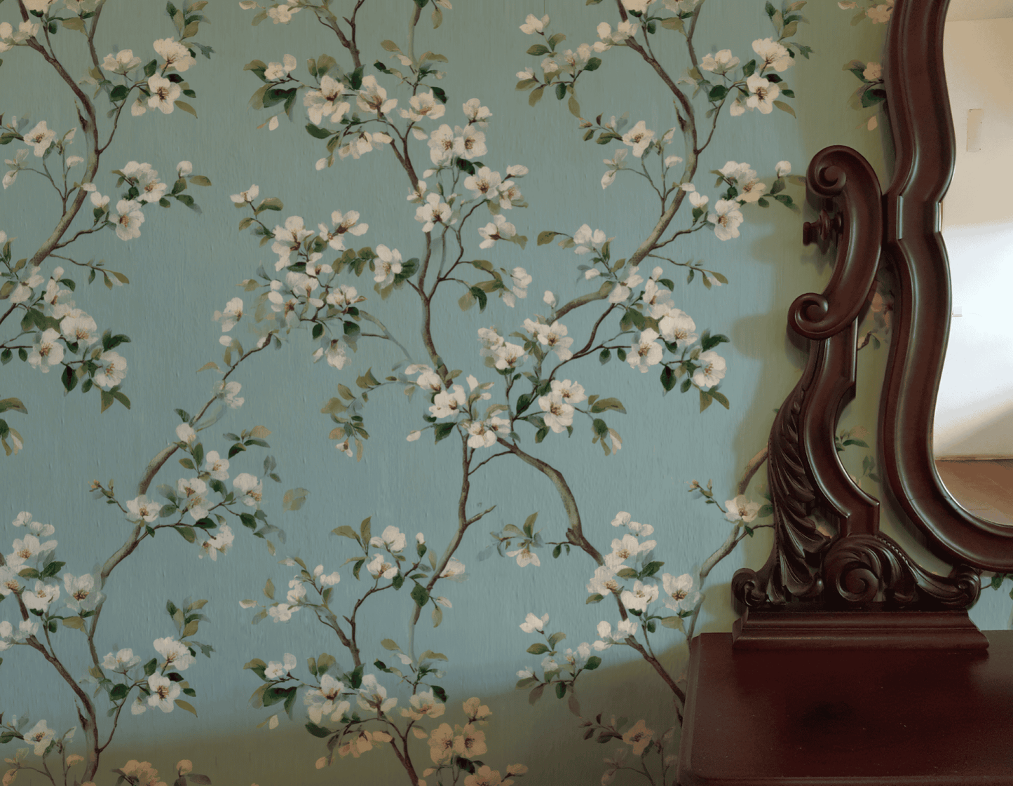 Blossom Dream Wallpaper features white blossoms on a light blue background, shown beside the ornately carved edge of a wooden mirror.