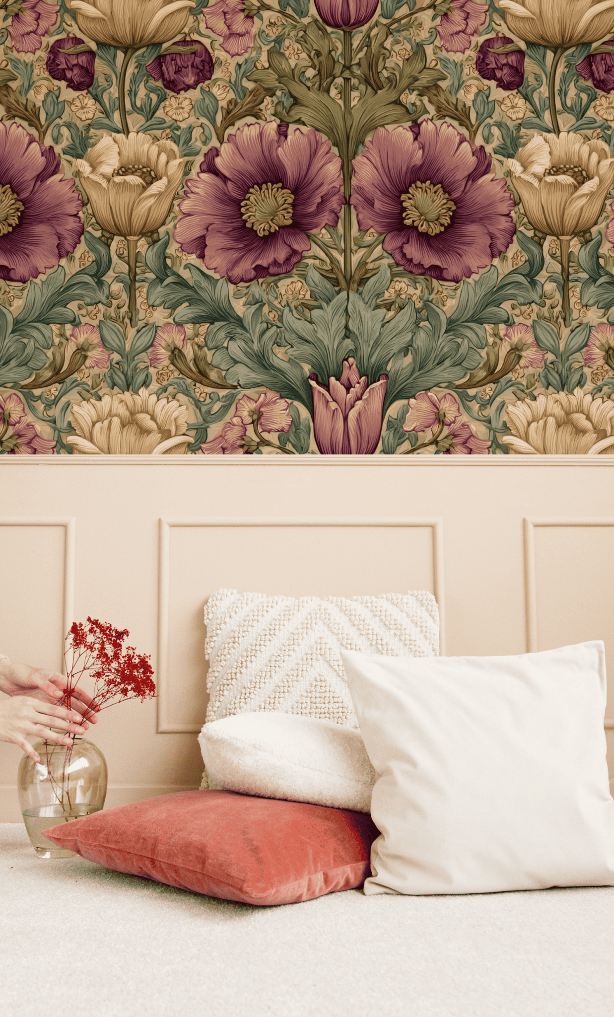 A hand sets a glass vase with red dried flowers and green foliage beside cream and coral pillows on a beige bench, in front of Victorian Blossom Wallpaper featuring large purple and yellow florals.