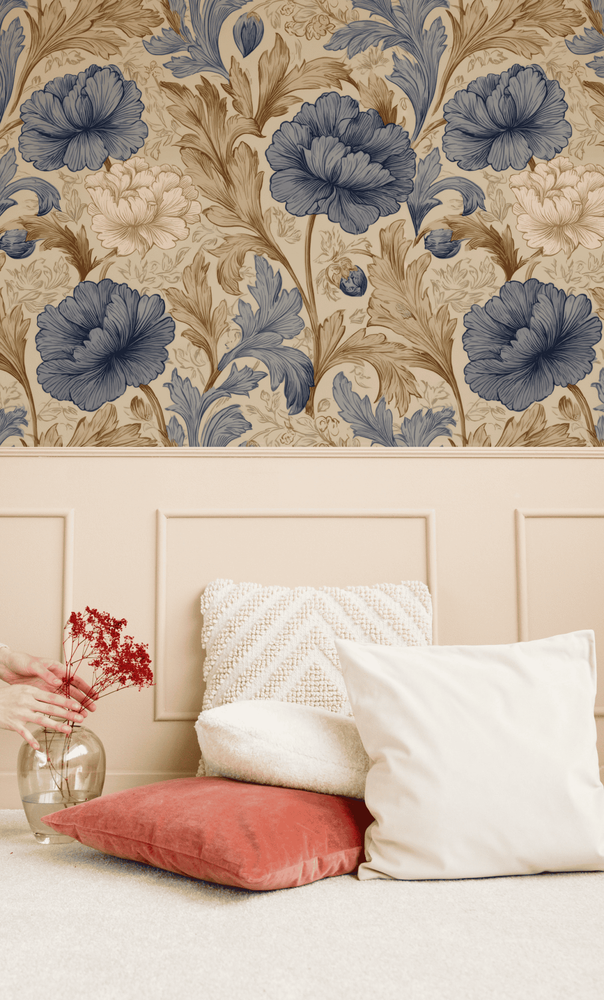 A hand sets a glass vase with red flowers on a beige surface beside three pillows, enhancing the timeless look against a paneled wall featuring Cassandra Bloom Wallpaper in elegant blue and beige florals.