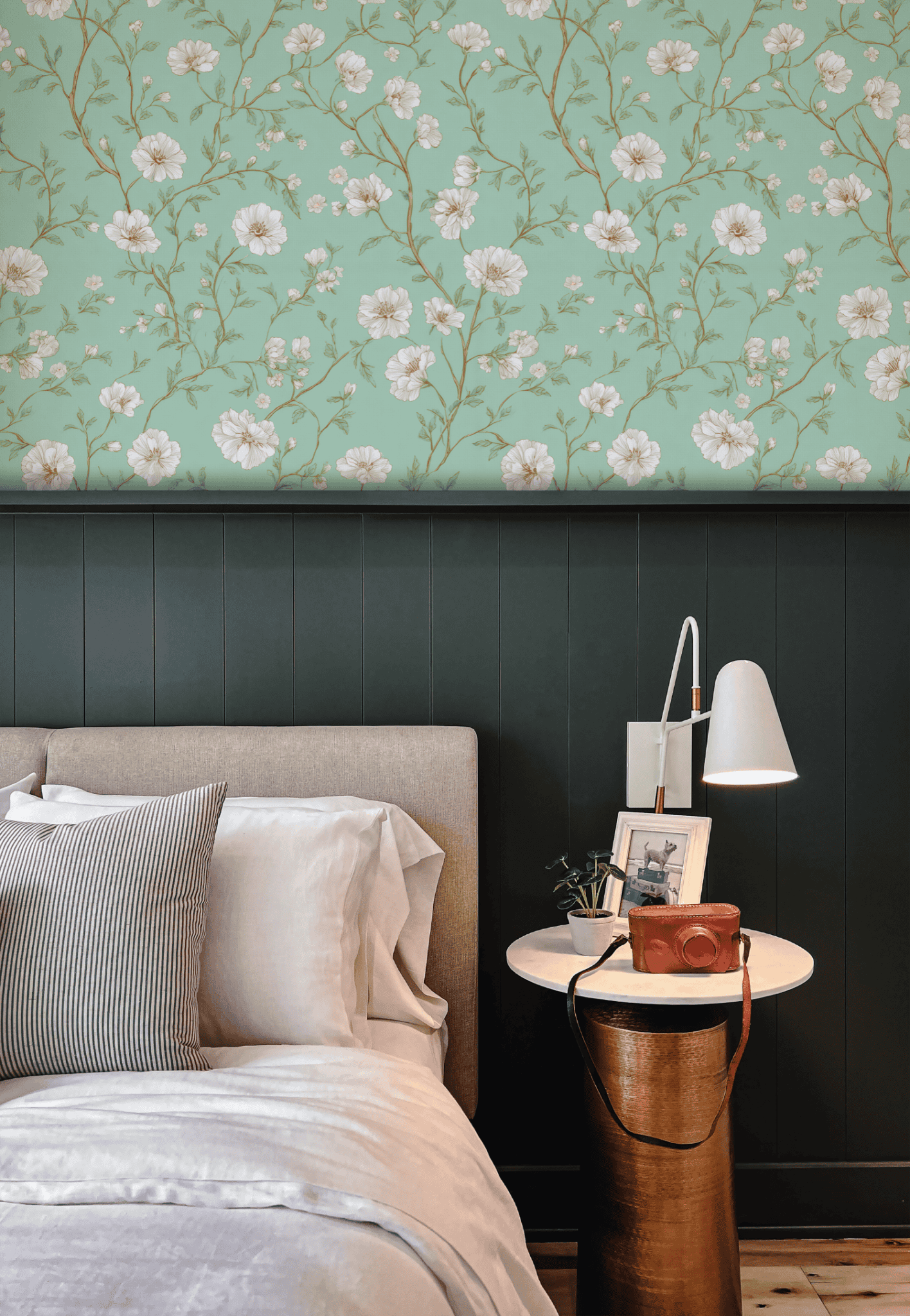 A neatly made bed with neutral bedding sits beside a round white nightstand topped with a lamp, books, and decor, set against Whispering Blooms Wallpaper featuring white flowers above rich dark green wainscoting.