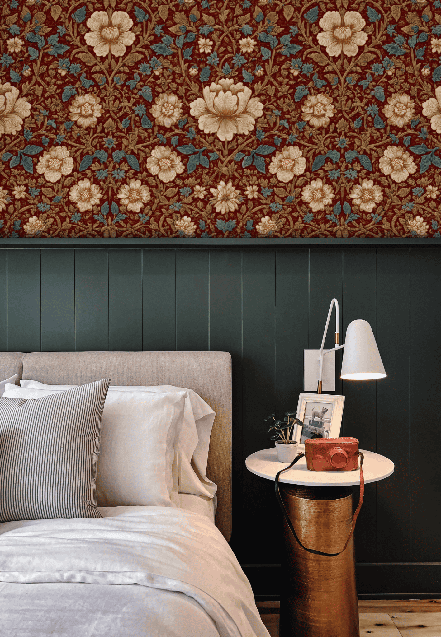 A neatly made bed with white linens and striped pillows sits by a round nightstand, lamp, and camera, set against a dark paneled wall topped with Victorian Blossom Wallpaper and classic decor above.