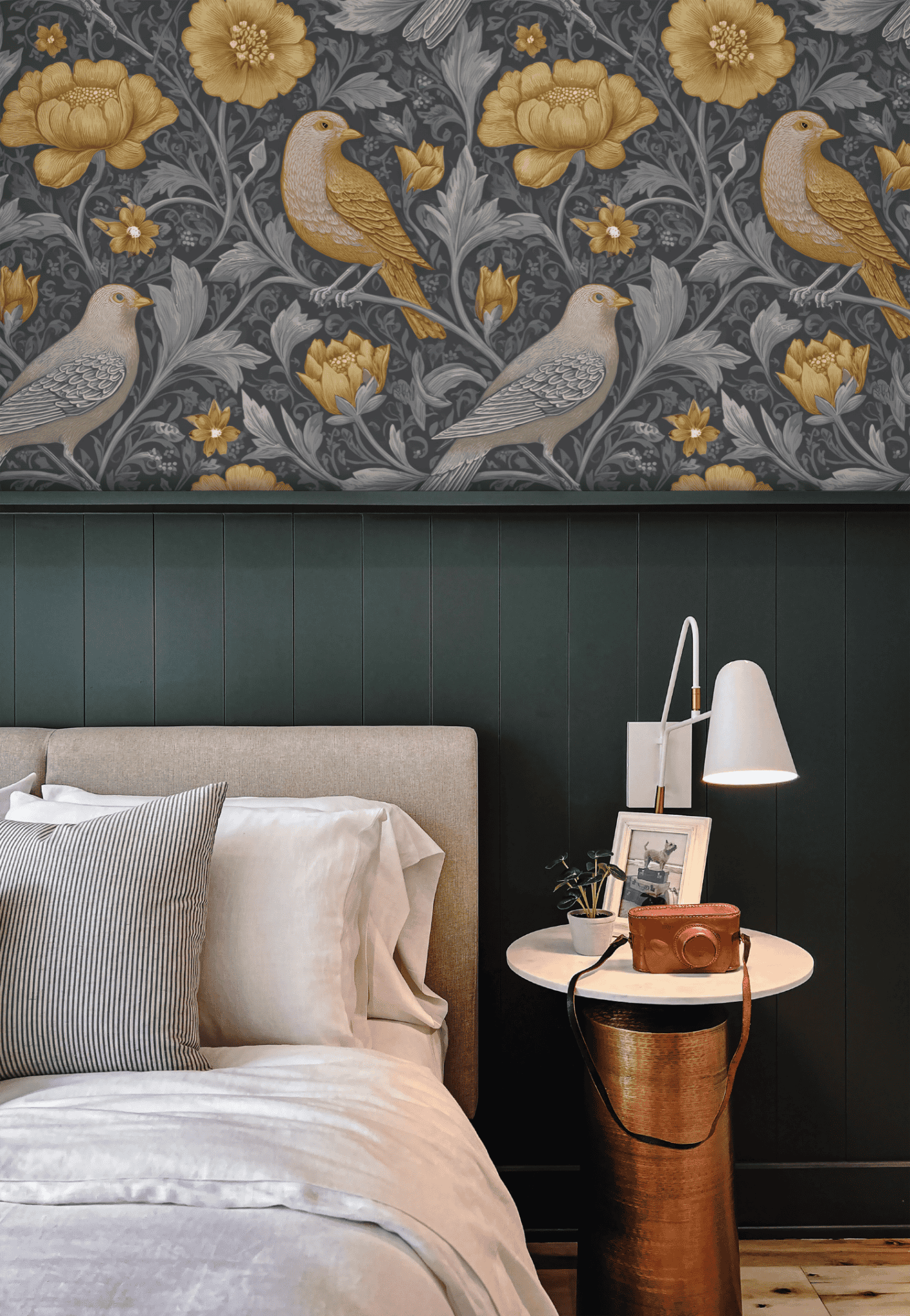 Create a sophisticated bedroom ambiance with the Golden Aviary Wallpaper, featuring golden and grey birds and blossoms on a dark background, paired with a bed, striped and white pillows, and a round side table with lamp and camera.