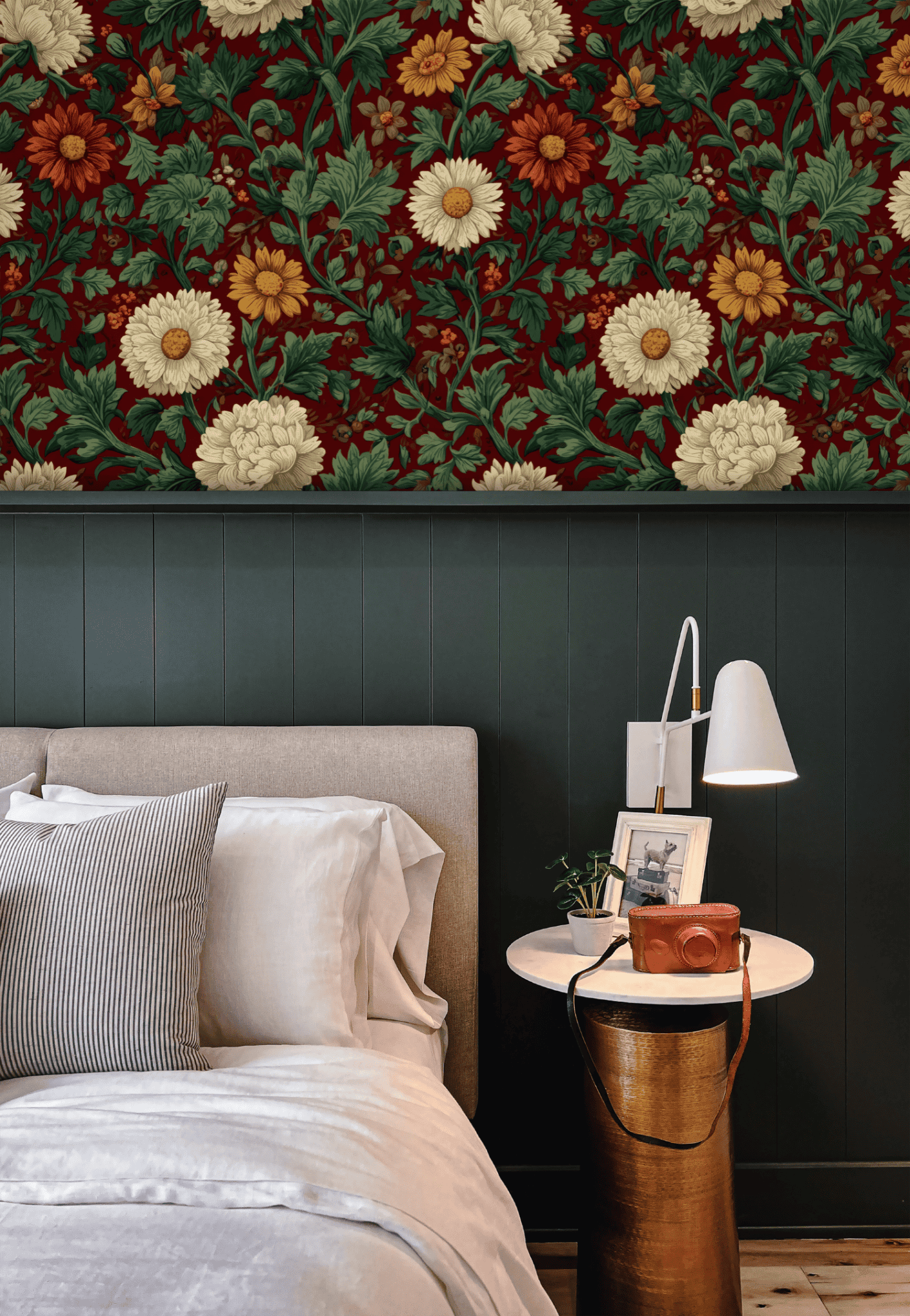 A modern bedroom features a beige bed, white linens, a striped pillow, and a round side table with lamp and camera. Crimson Blossoms Wallpaper above dark green wall paneling brings vintage charm to the space.