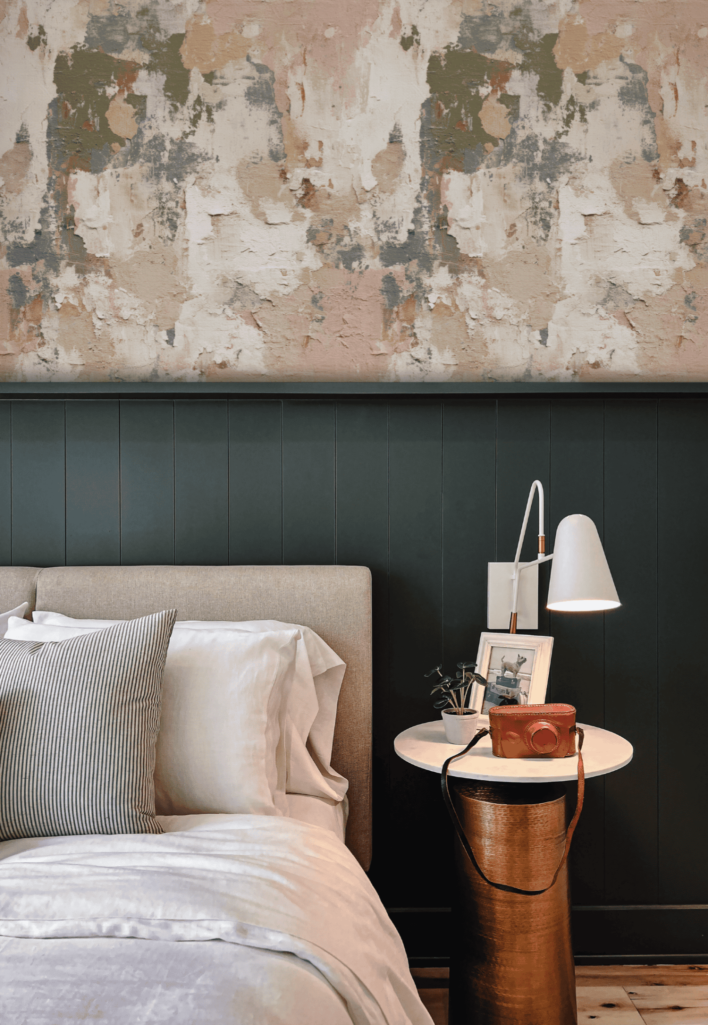 A modern bedroom with a beige upholstered bed and striped pillow exudes elegance, featuring a round side table with decor, set against a dark green paneled wall and Rustic Plaster textured abstract wallpaper.