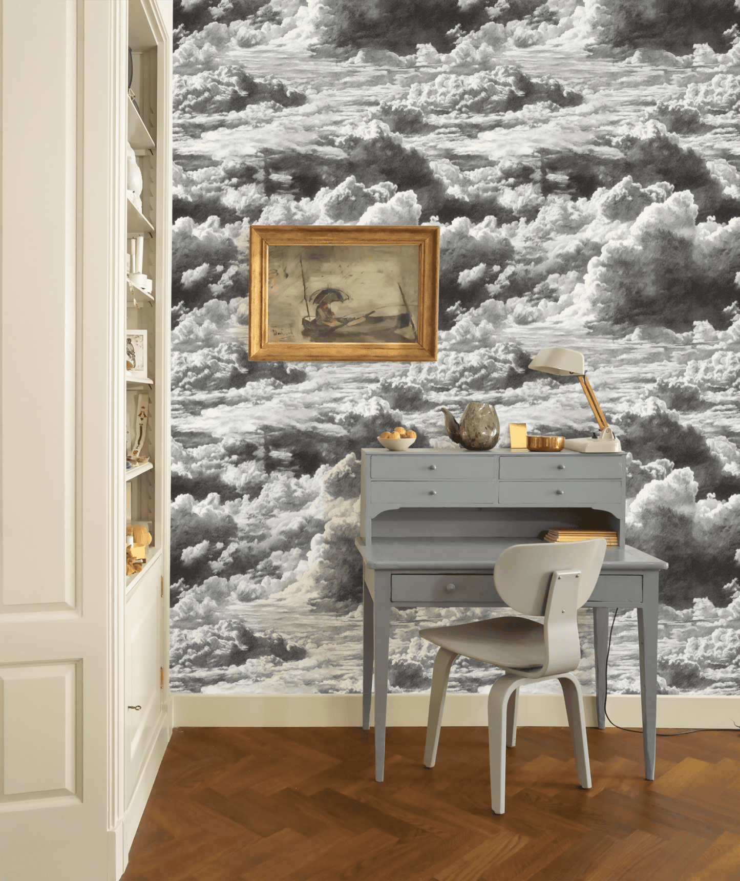 A small gray desk with a matching chair and lamp set against Nimbus Reverie Wallpaper, swirling in monochrome clouds, with a framed painting above—a perfect spot for serene daydreaming.