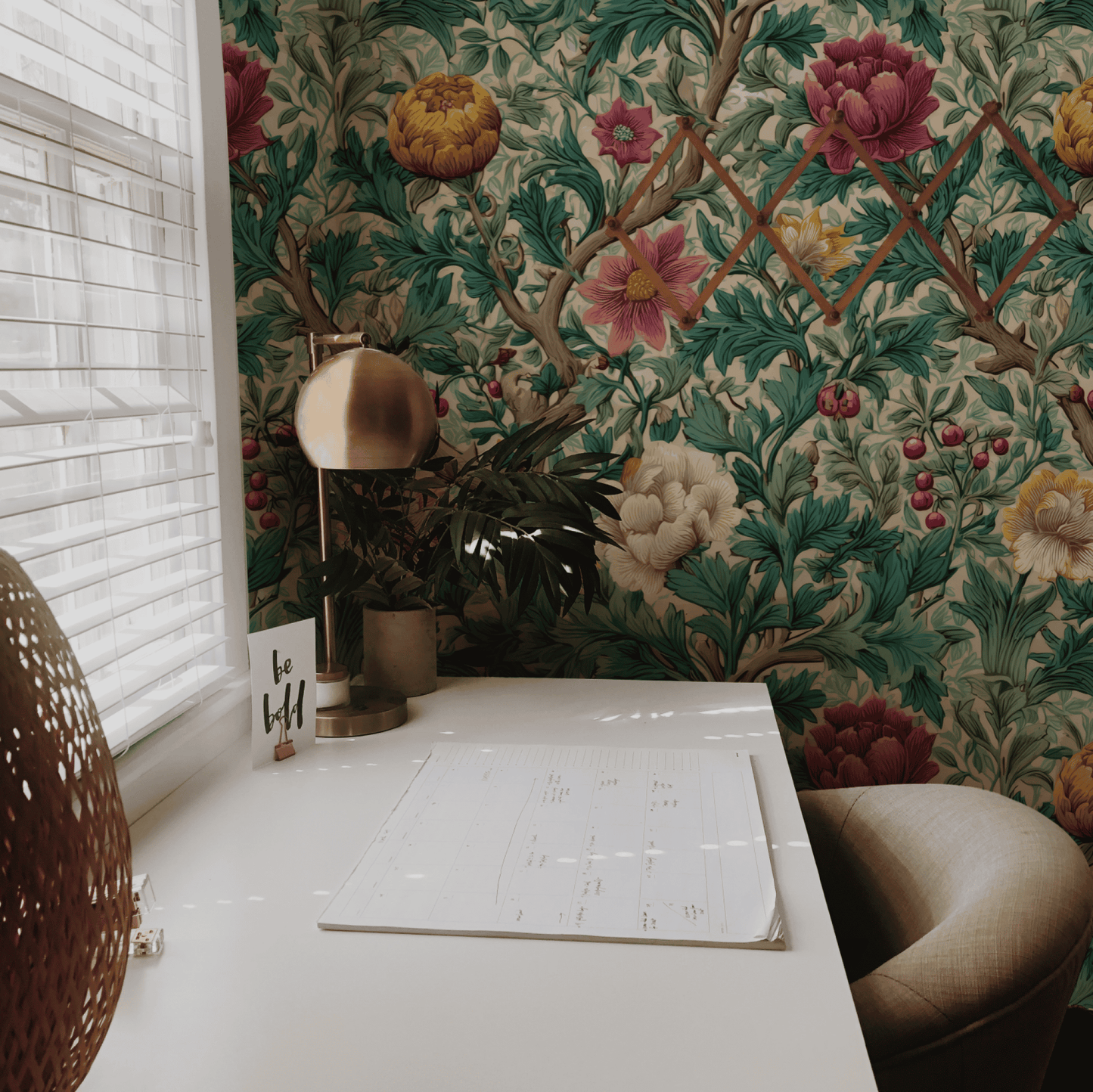 A white desk with a calendar, lamp, and chair sits by a window; the wall behind features Botanical Whimsy Wallpaper with green leaves and pink, yellow, and red flowers for a vibrant botanical garden feel.