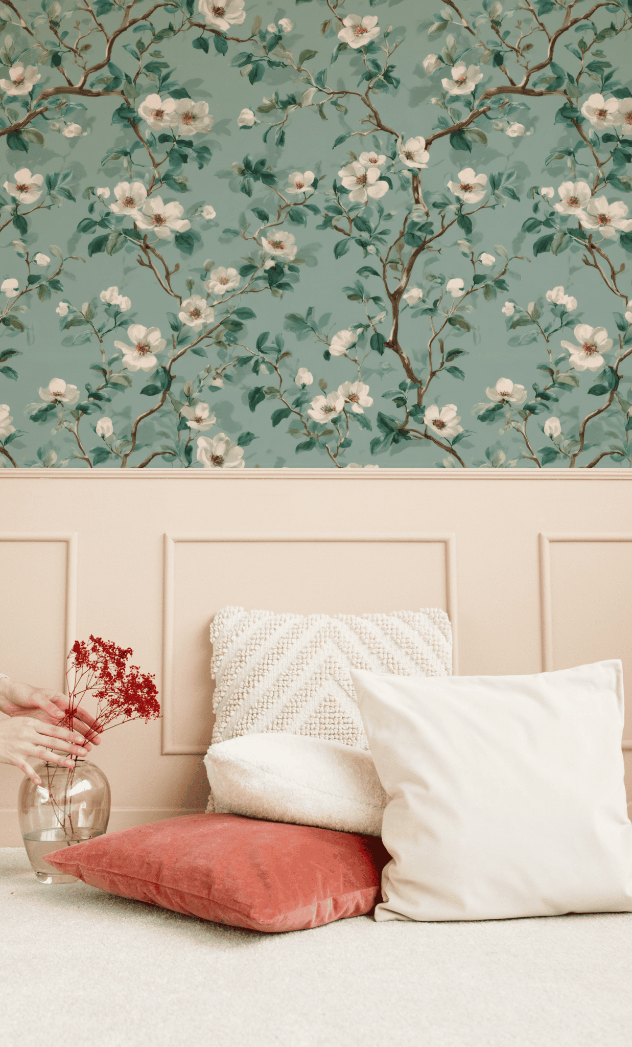 Two pillows rest on a carpeted floor against a beige paneled wall; Blossom Whisper Wallpaper with verdant leaves and a glass vase with red dried flowers are also visible.
