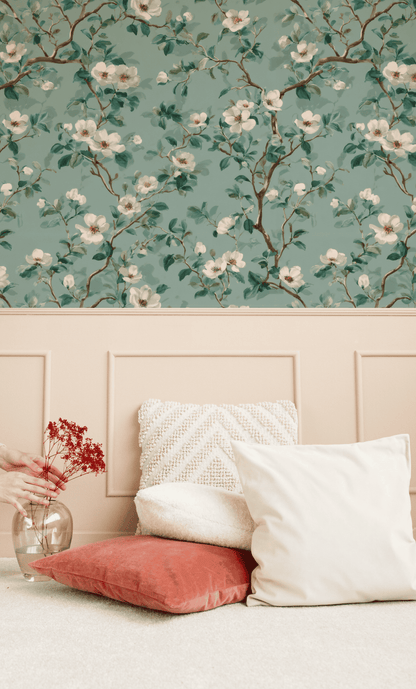 Two pillows rest on a carpeted floor against a beige paneled wall; Blossom Whisper Wallpaper with verdant leaves and a glass vase with red dried flowers are also visible.