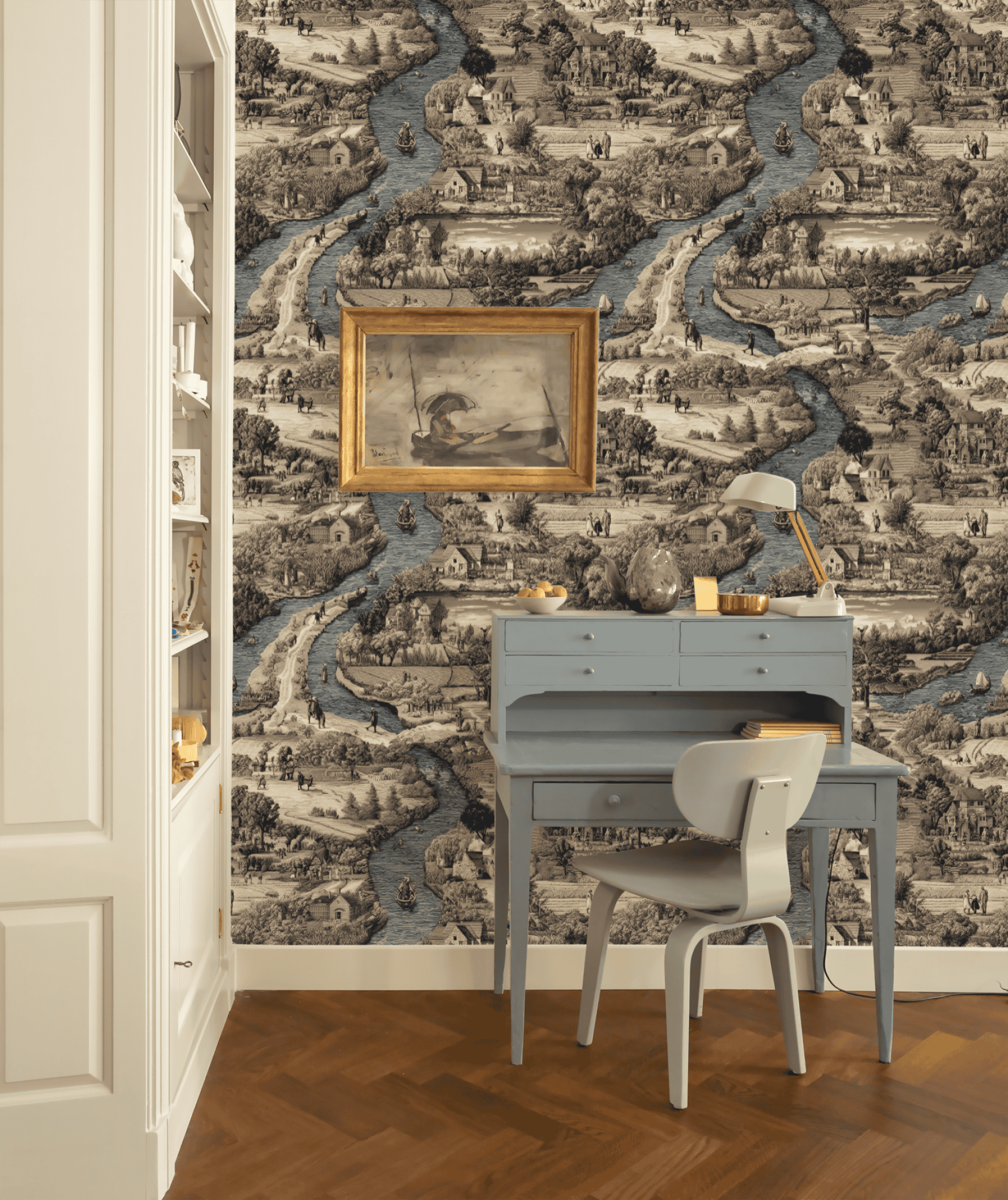 A small blue desk with chair and lamp sits before Serene Pastoral Wallpaper and a framed vintage landscape, next to built-in shelves in a cozy home office.