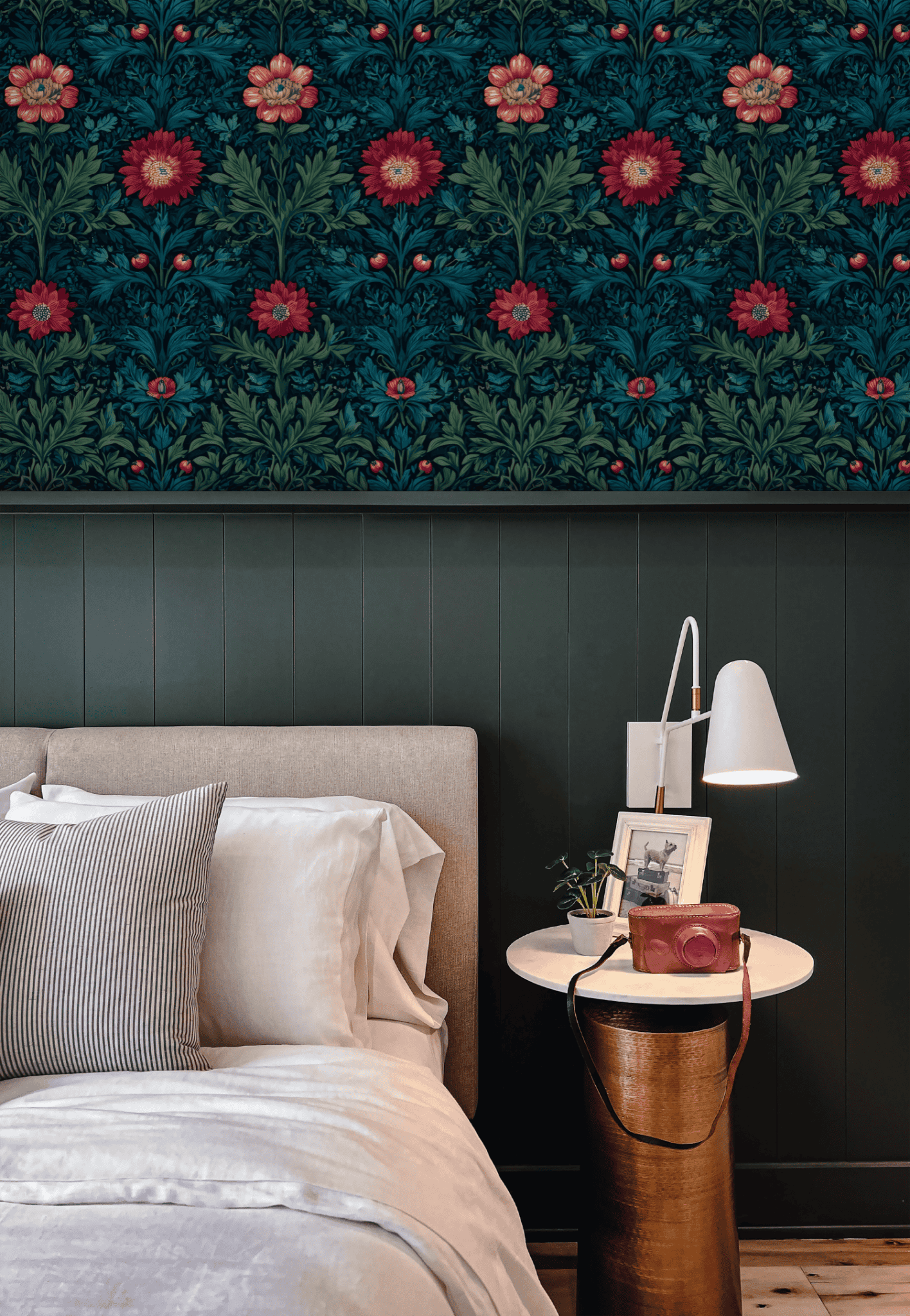 A modern bedroom features a beige bed, striped pillows, round white side table, white lamp, and elegant Crimson Blooms Wallpaper with red flowers above dark wainscoting for a sophisticated decor touch.