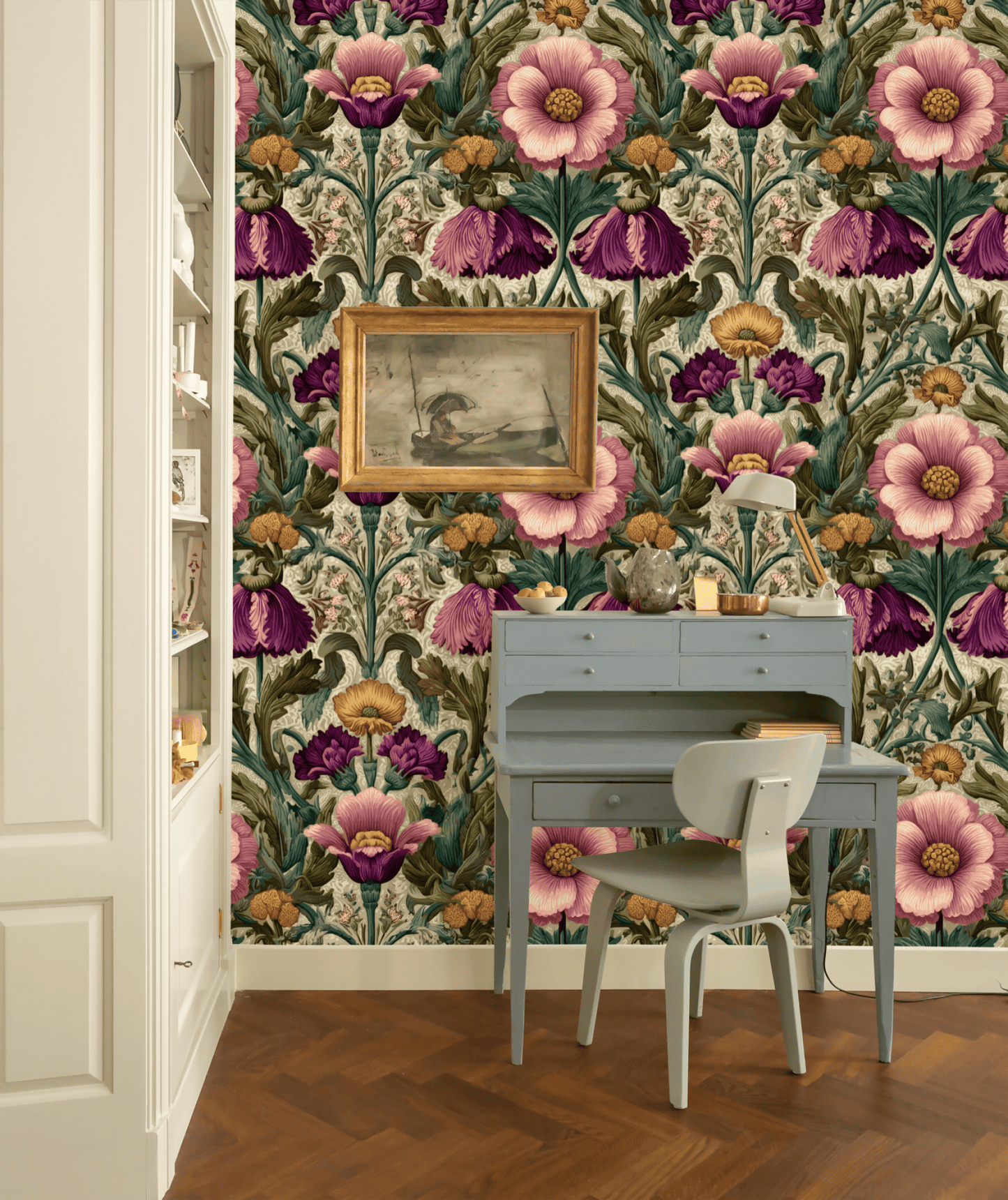 A small gray desk and chair sit against a wall of Blossom Rhapsody Wallpaper in bold pink, purple, and green florals; a framed painting and decorative items add classic elegance on and above the desk.