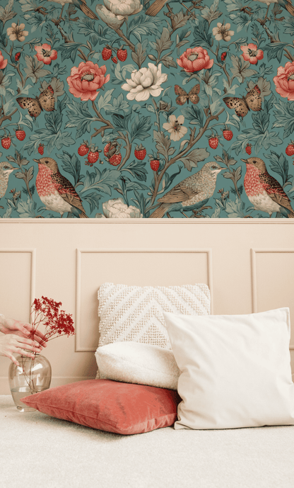 A hand holds a vase with red flowers beside cream and coral cushions. The wall behind features Garden Serenade Wallpaper, adorned with flowers and birds above beige paneling.
