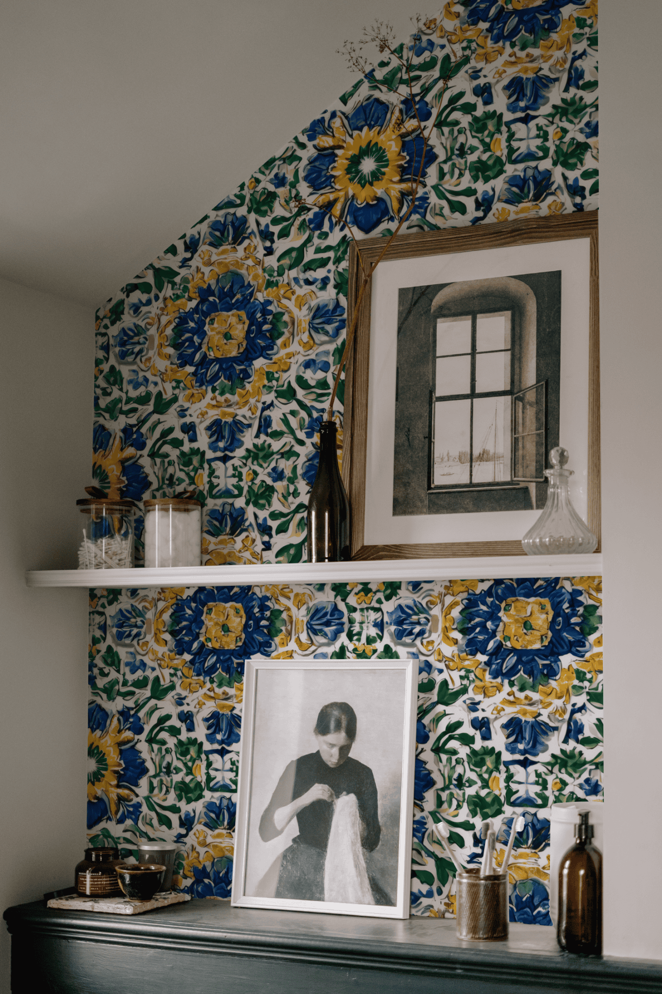 A wall covered in Mediterranean Bloom Wallpaper displays two shelves with framed art, glass bottles, and small decorative items.