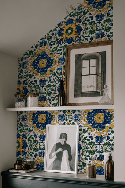 A wall covered in Mediterranean Bloom Wallpaper displays two shelves with framed art, glass bottles, and small decorative items.