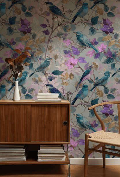 A wooden sideboard with stacked books and a vase stands against a wall covered in Enchanted Aviary Wallpaper, next to a wooden chair with a woven seat.