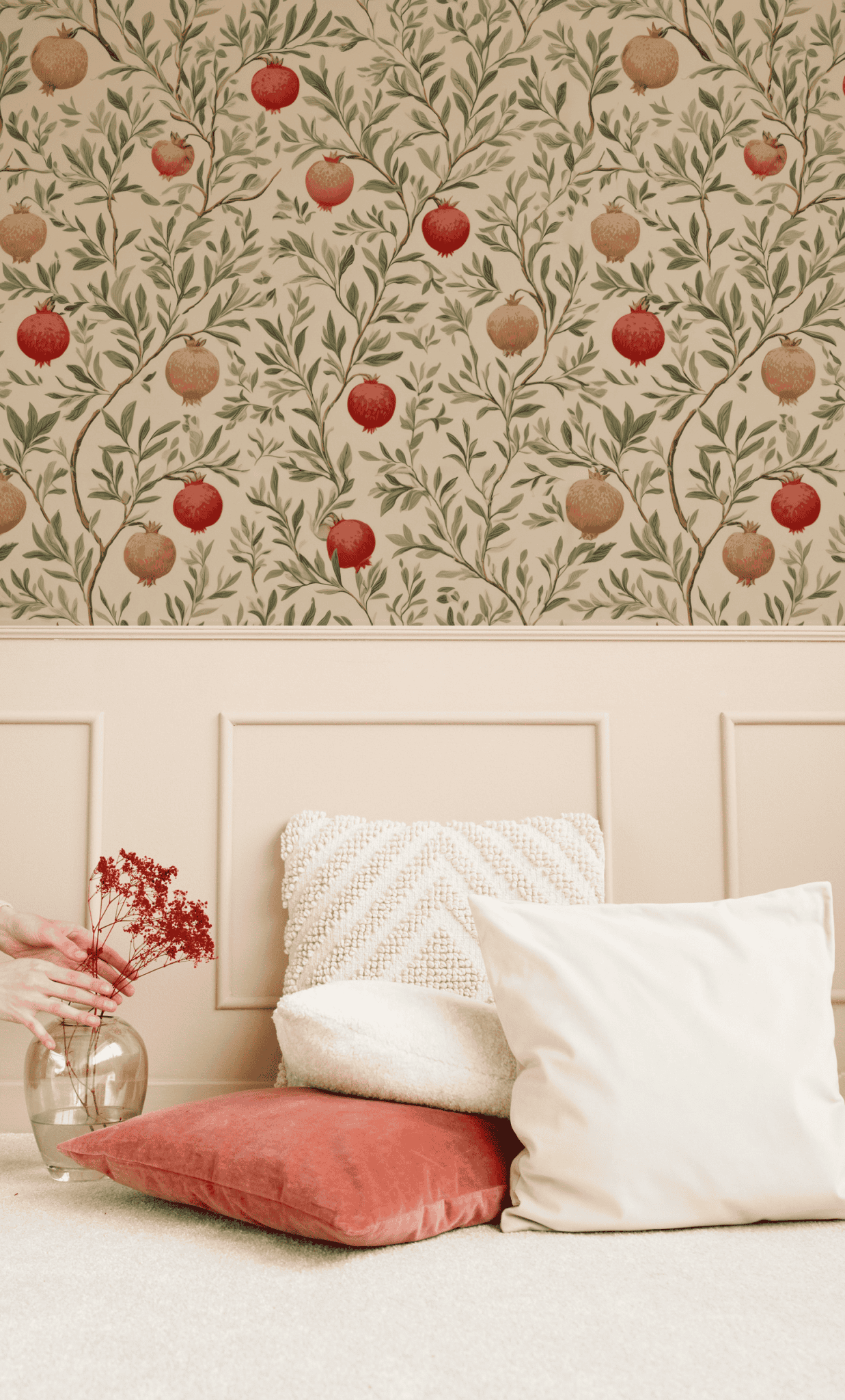 A hand holds a glass vase with red dried flowers and leafy branches on the floor beside white and coral pillows, set against a beige wall decorated with Pomegranate Grove Wallpaper.