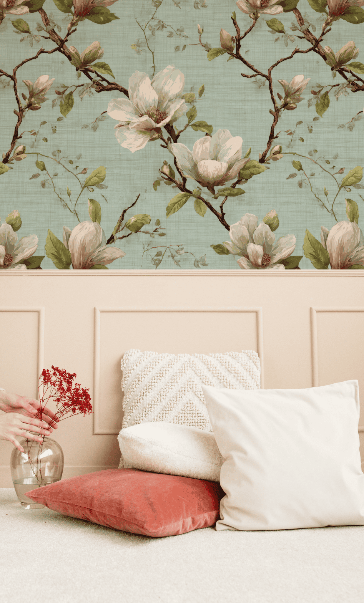 A hand arranges red dried flowers in a glass vase on a beige surface beside white and coral pillows, with Magnolia Dream Wallpaper and magnolia blossoms creating a serene atmosphere.