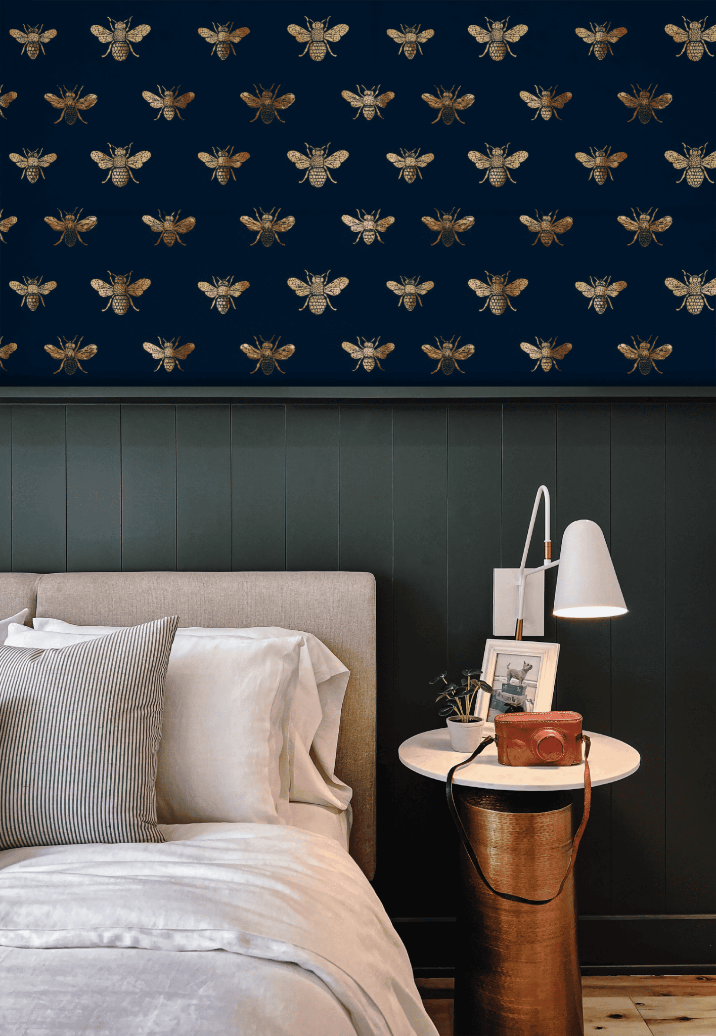 A bedroom showcases Golden Hive Wallpaper—a dark navy wall with gold bees—paired with a beige bed, white bedding, and a round side table holding a lamp and camera to create a luxurious ambiance.