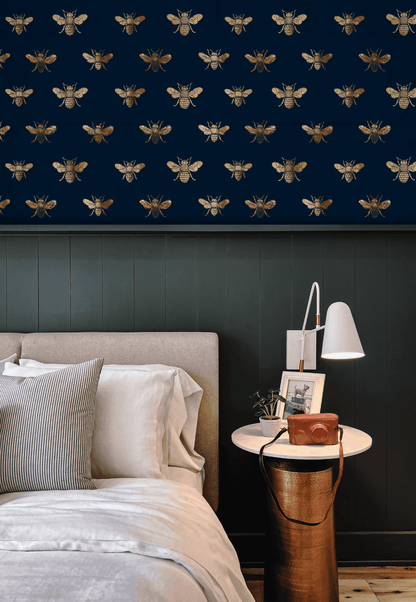 A bedroom showcases Golden Hive Wallpaper—a dark navy wall with gold bees—paired with a beige bed, white bedding, and a round side table holding a lamp and camera to create a luxurious ambiance.