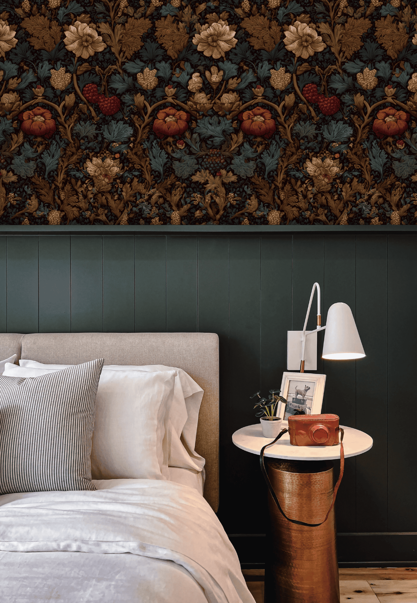 A neatly made bed with beige bedding sits by a white round side table holding a lamp, books, and a camera. The wall behind features Autumn Grove Wallpaper with delicate blossoms above green paneling.