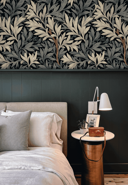 A modern bedroom showcases a beige upholstered bed, white bedding, a round nightstand with a lamp, and Eclipse Flora Wallpaper featuring a dark botanical floral design above green paneling for a sophisticated ambiance.