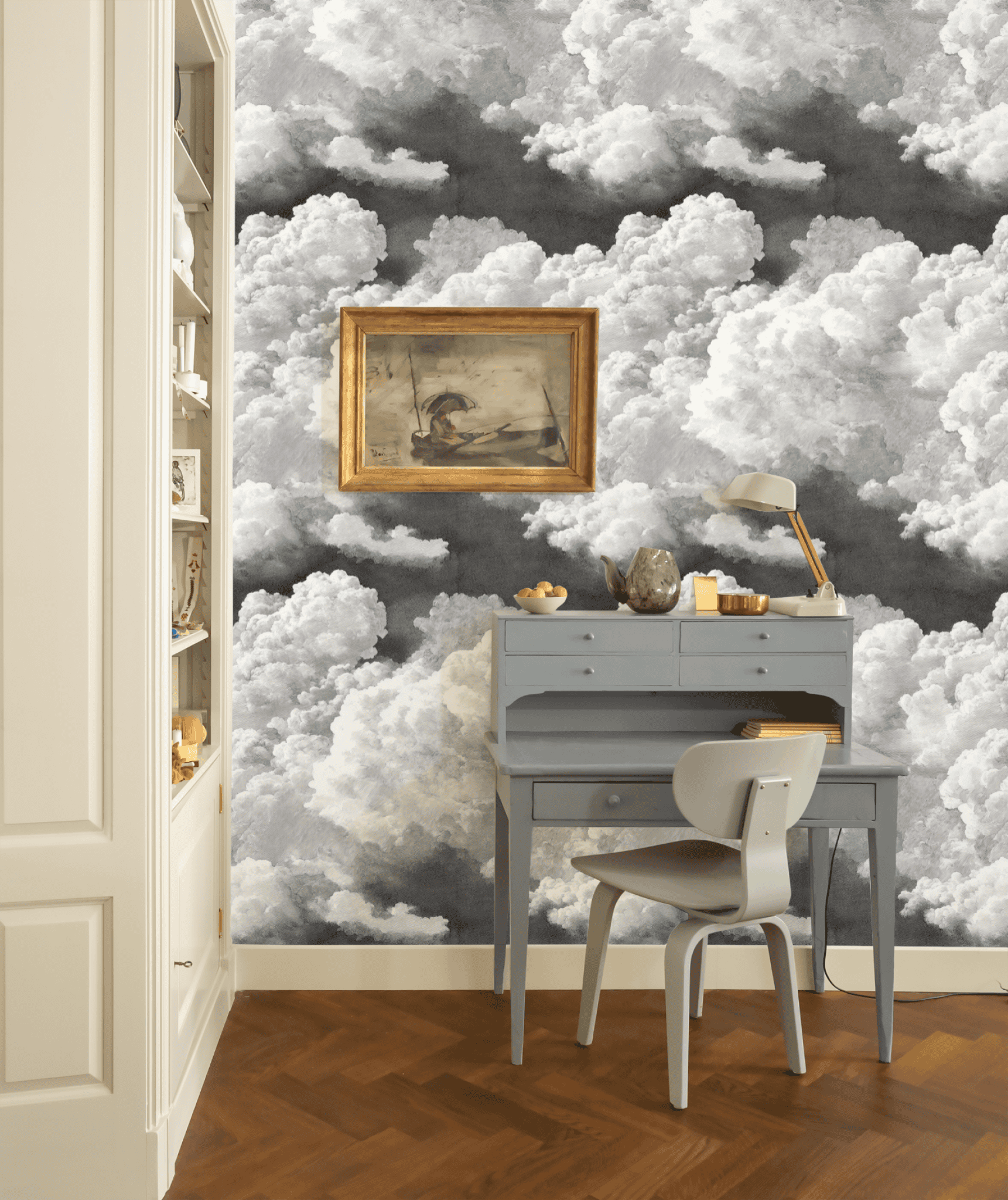 A small gray desk with a chair sits against a wall covered in Cloud Drift Wallpaper, creating a dreamy ambiance. A framed painting and desk lamp are on the desk, while shelves are visible to the left.