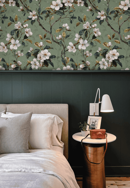 A modern bedroom with a light upholstered bed, striped pillows, a round side table holding books, a camera, and a white lamp, set against elegant green floral Blossom Serenity Wallpaper and a dark paneled wall.