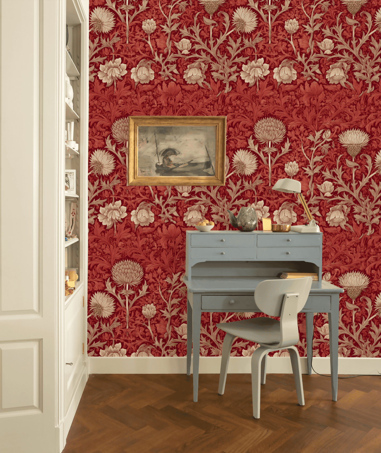 A small blue writing desk and chair sit against a backdrop of Crimson Elegance Wallpaper with red floral patterns, accented by a framed painting and decor above. Bookshelves are visible to the left.