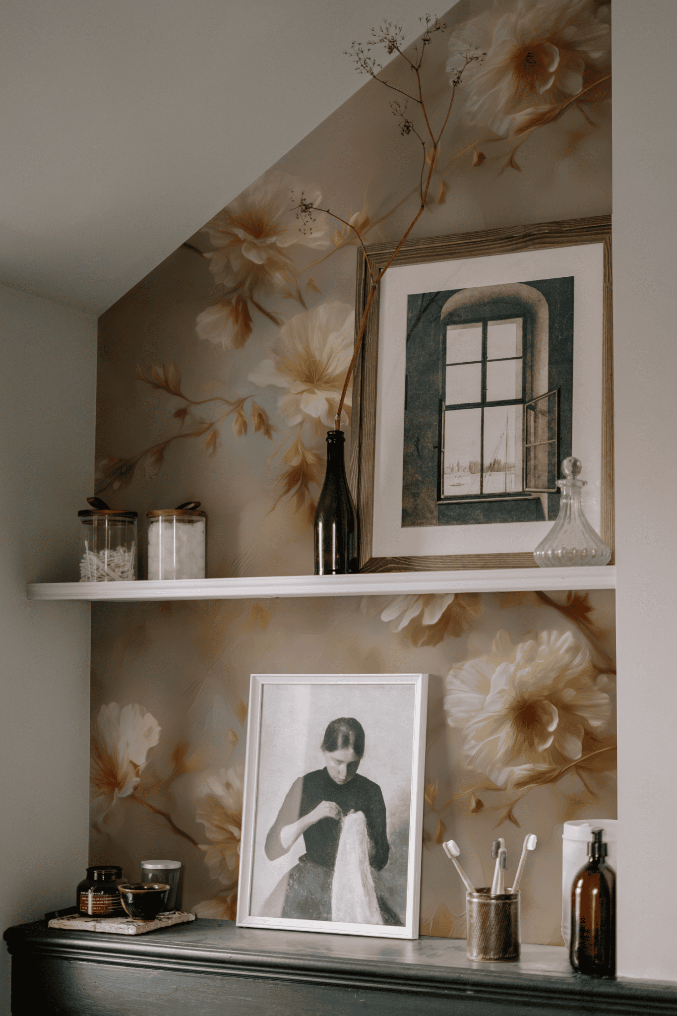 Two shelves with framed photos, glass jars, and bottles are mounted on a wall adorned with Whispering Blossom Wallpaper above a dark cabinet holding brushes and toiletries, creating an atmosphere of timeless elegance.