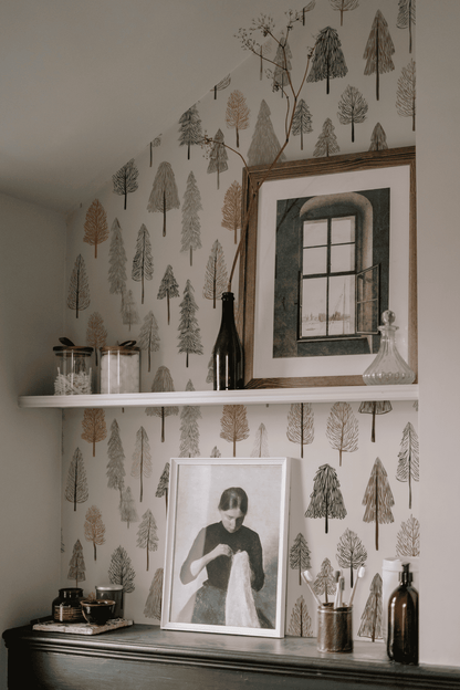 A shelf on a wall covered in Whispering Pines Wallpaper holds bottles, jars, and a framed window photo. Below, a black-and-white portrait of a woman sits on a dark cabinet, bringing woodland charm to the space.