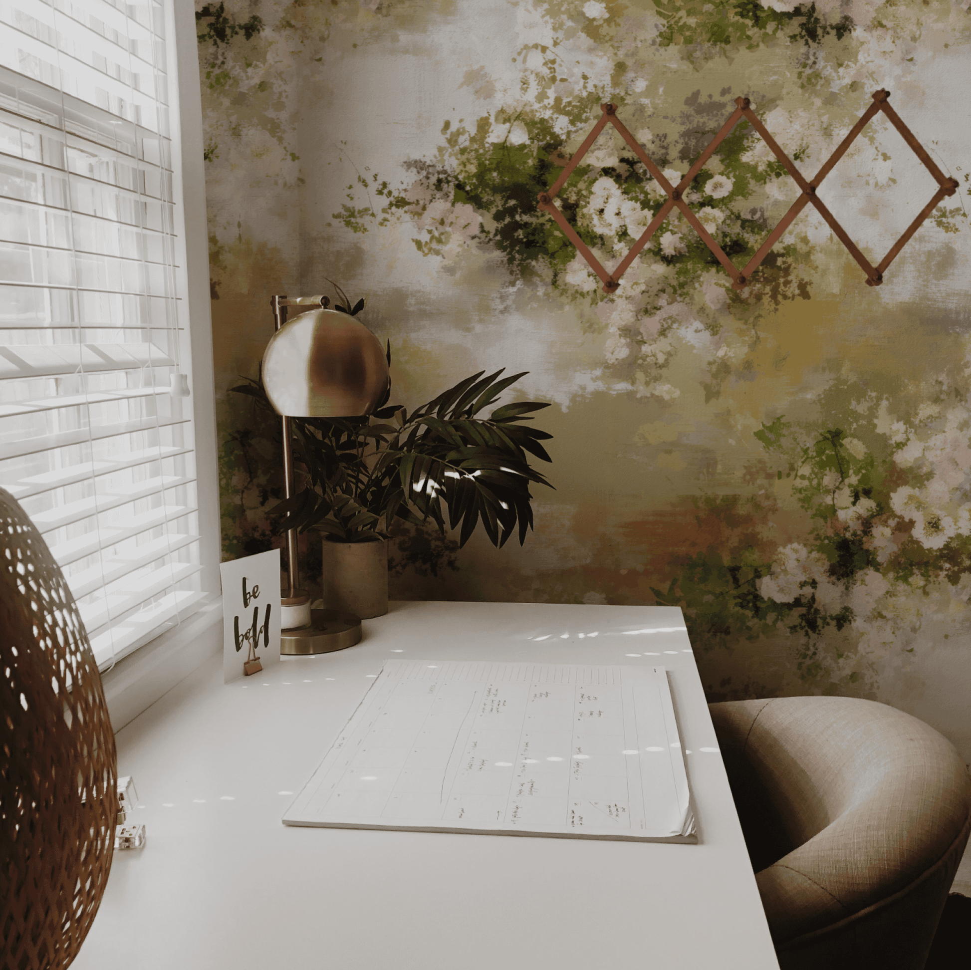A white desk with a calendar, lamp, plant, and “be bold” card sits by a window with blinds. Whispering Petals Wallpaper and subtle hues throughout create a romantic ambiance, complemented by a wooden wall rack in the background.