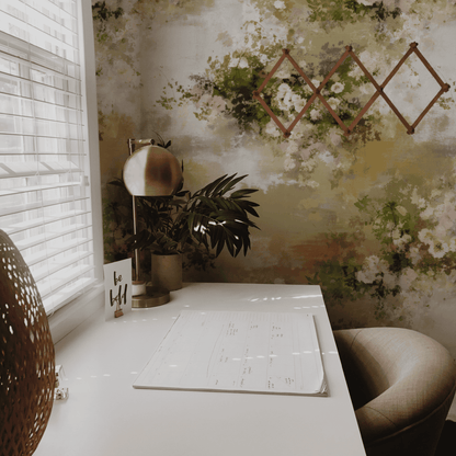 A white desk with a calendar, lamp, plant, and “be bold” card sits by a window with blinds. Whispering Petals Wallpaper and subtle hues throughout create a romantic ambiance, complemented by a wooden wall rack in the background.