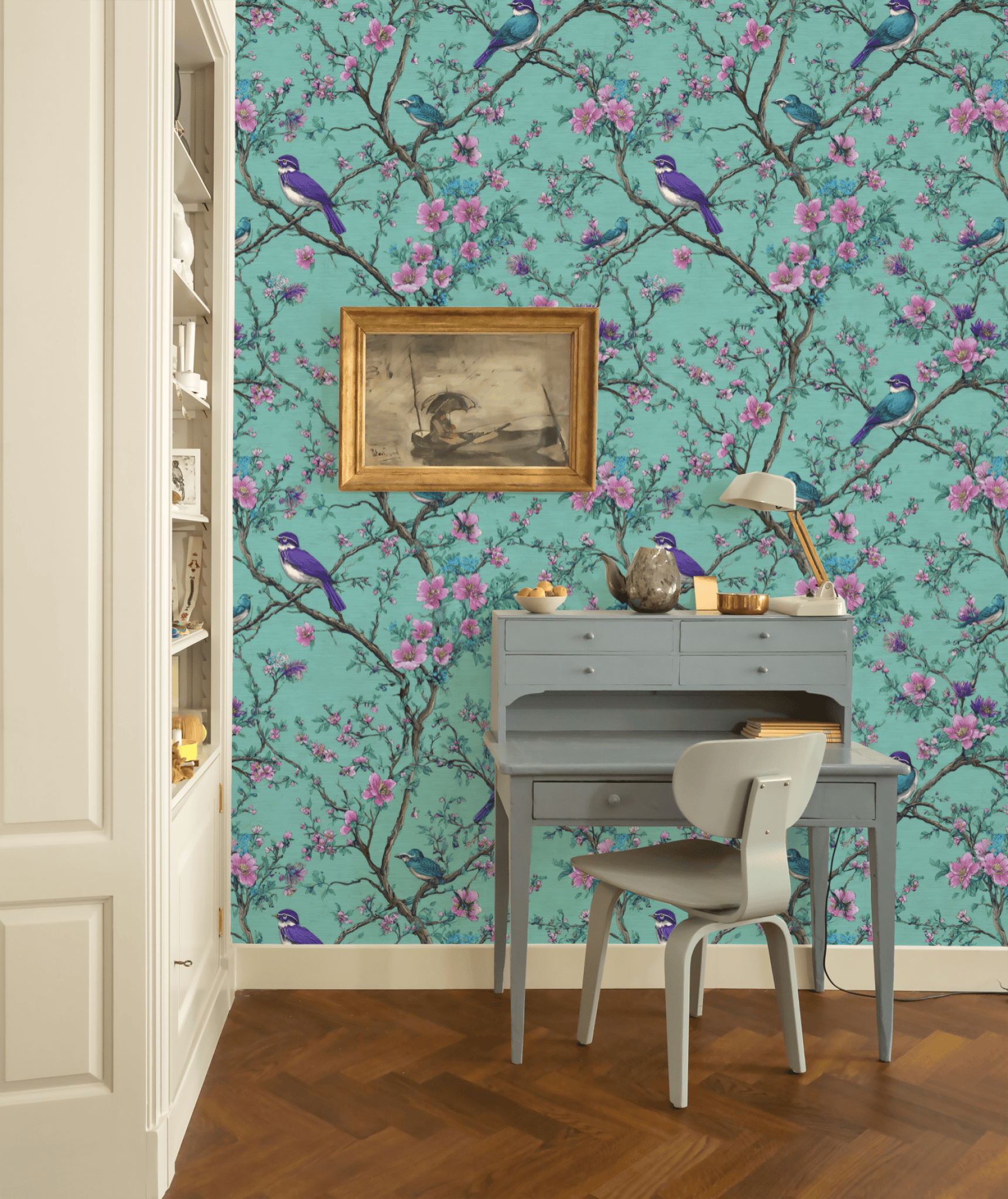 A small blue writing desk with a lamp, chair, and tea set sits against a wall decorated with Blossom Serenity Wallpaper, featuring vibrant birds and pink blossoms. A framed painting hangs above, and built-in shelves are partially visible on the left.